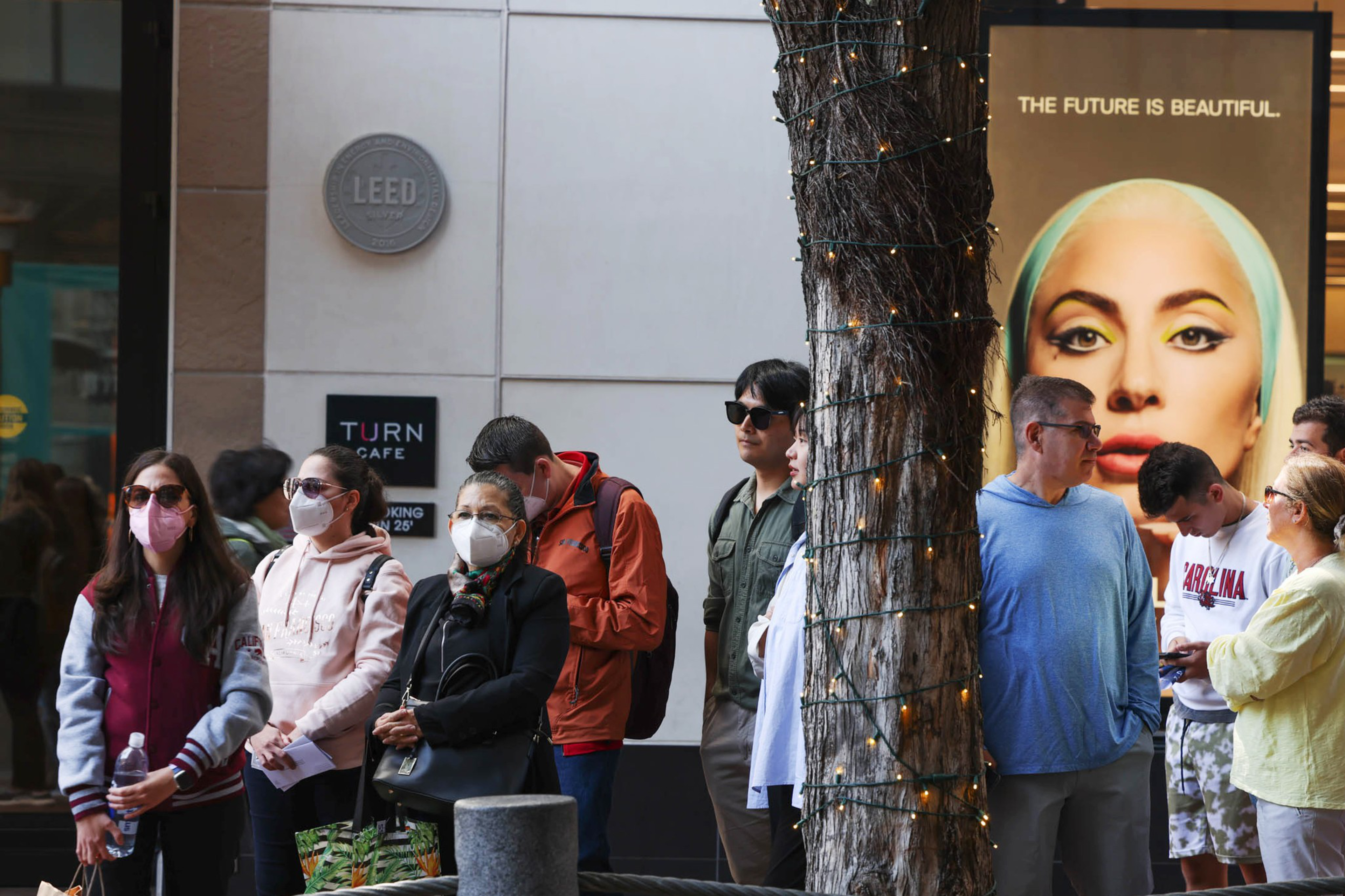 A diverse group of people, some wearing masks, stand near a tree wrapped in string lights next to a large billboard featuring a woman’s face with the text "THE FUTURE IS BEAUTIFUL."