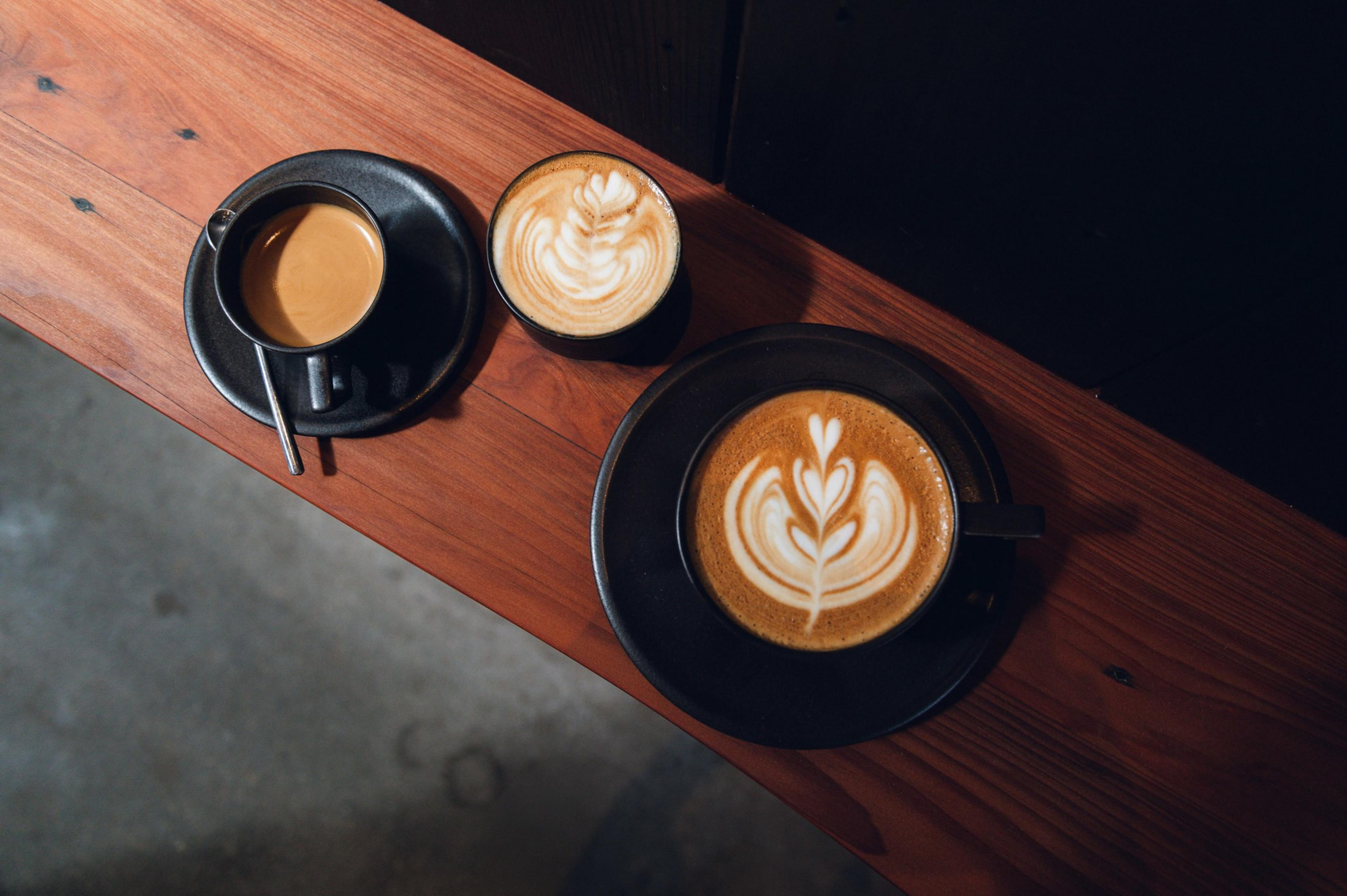 Three coffee cups on a wooden surface, two with latte art of leaves and one plain espresso in black cups and saucers with a spoon.