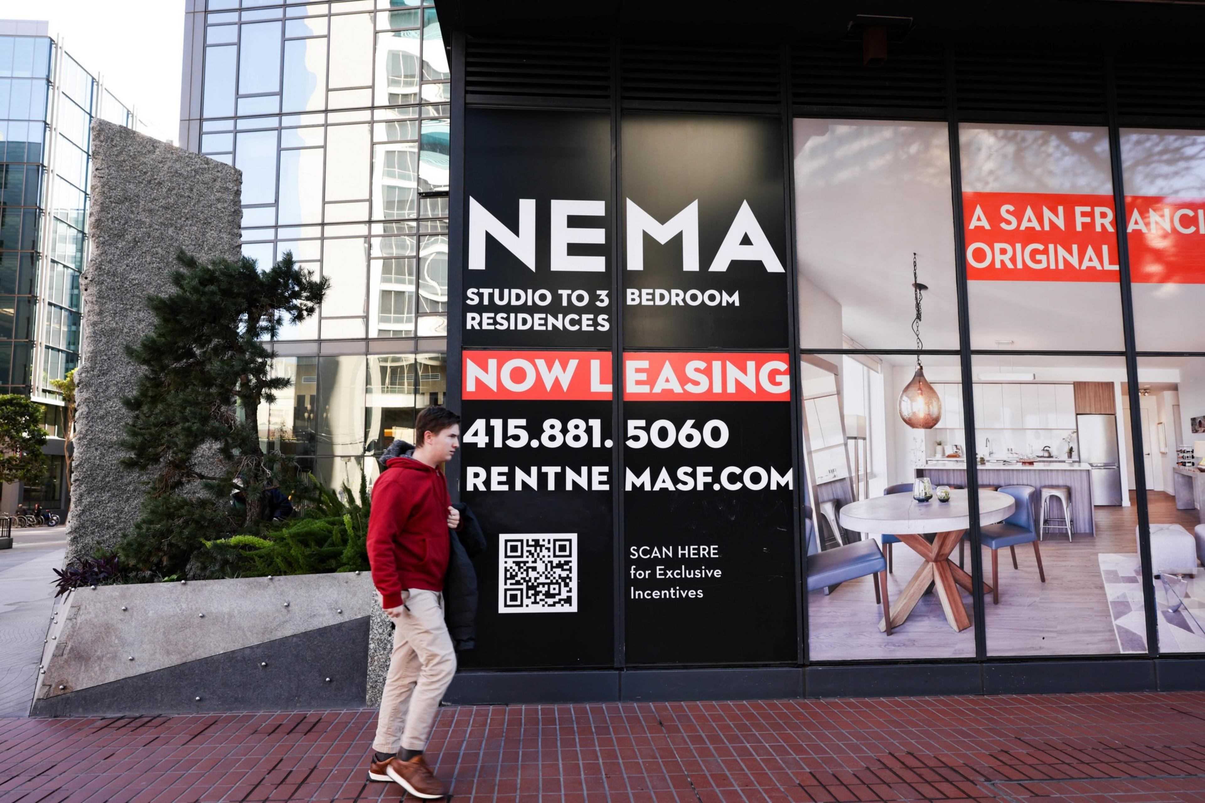 A person in a red jacket walks past a building with large signage for apartment leasing, featuring a modern interior image, contact info, and a QR code.