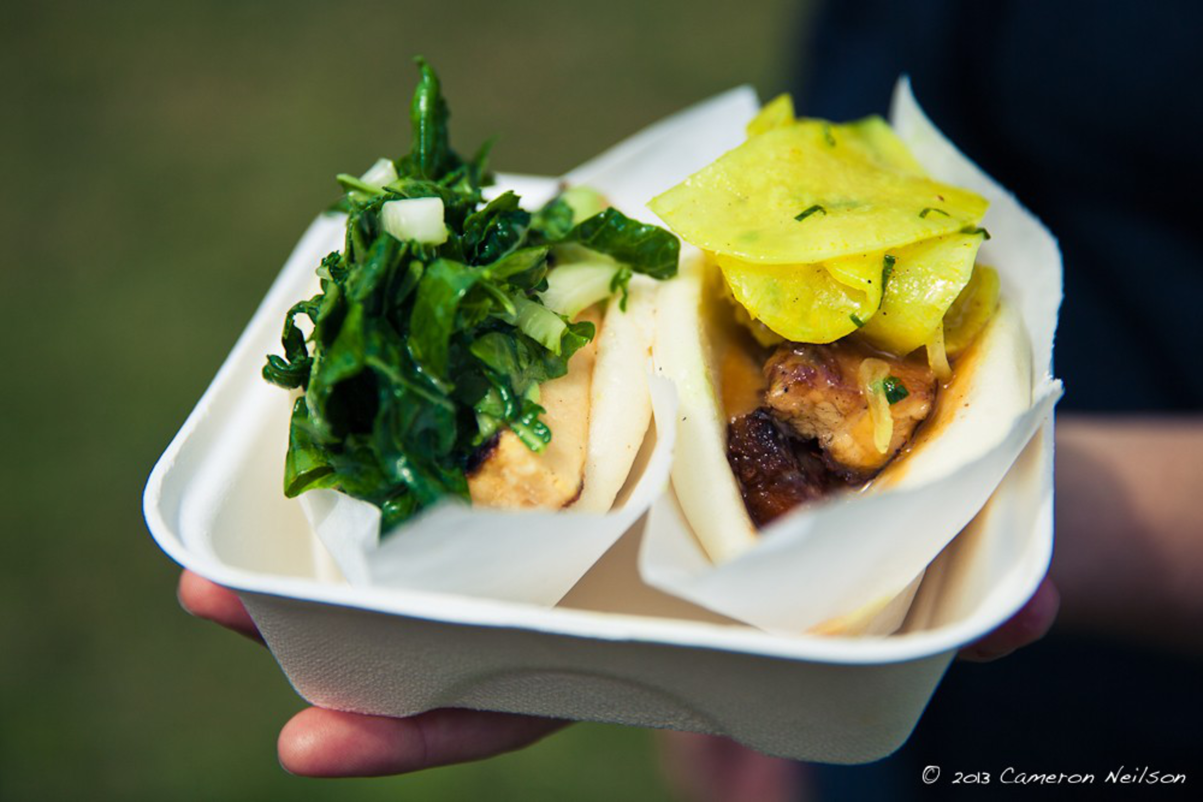 Two steamed buns filled with grilled meat, one topped with sautéed greens and onions, the other with pickled yellow radish and herbs.
