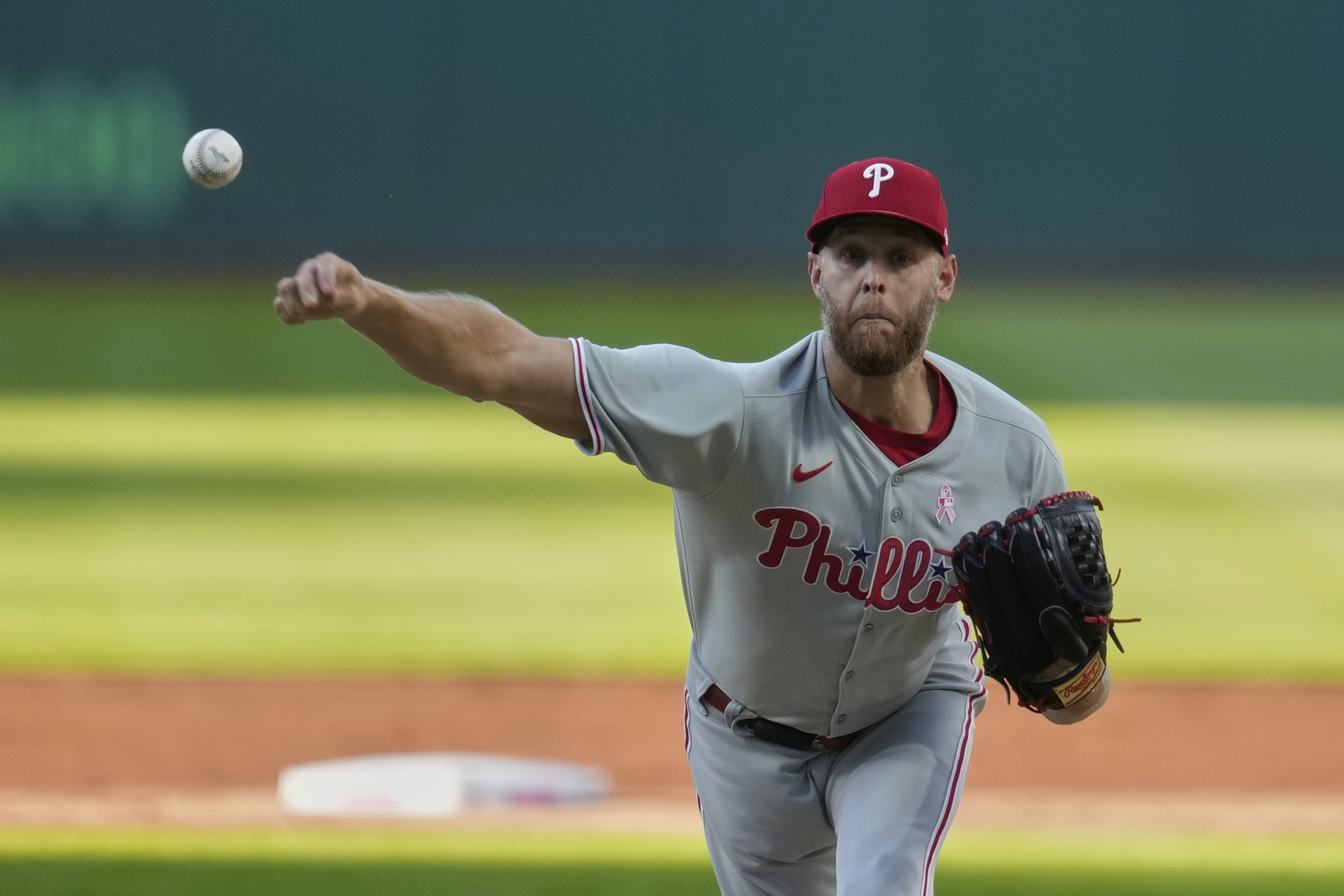 A baseball player in a Phillies gray uniform pitches a ball. He wears a red cap with a &quot;P&quot; logo, and the focus is on the ball mid-flight.