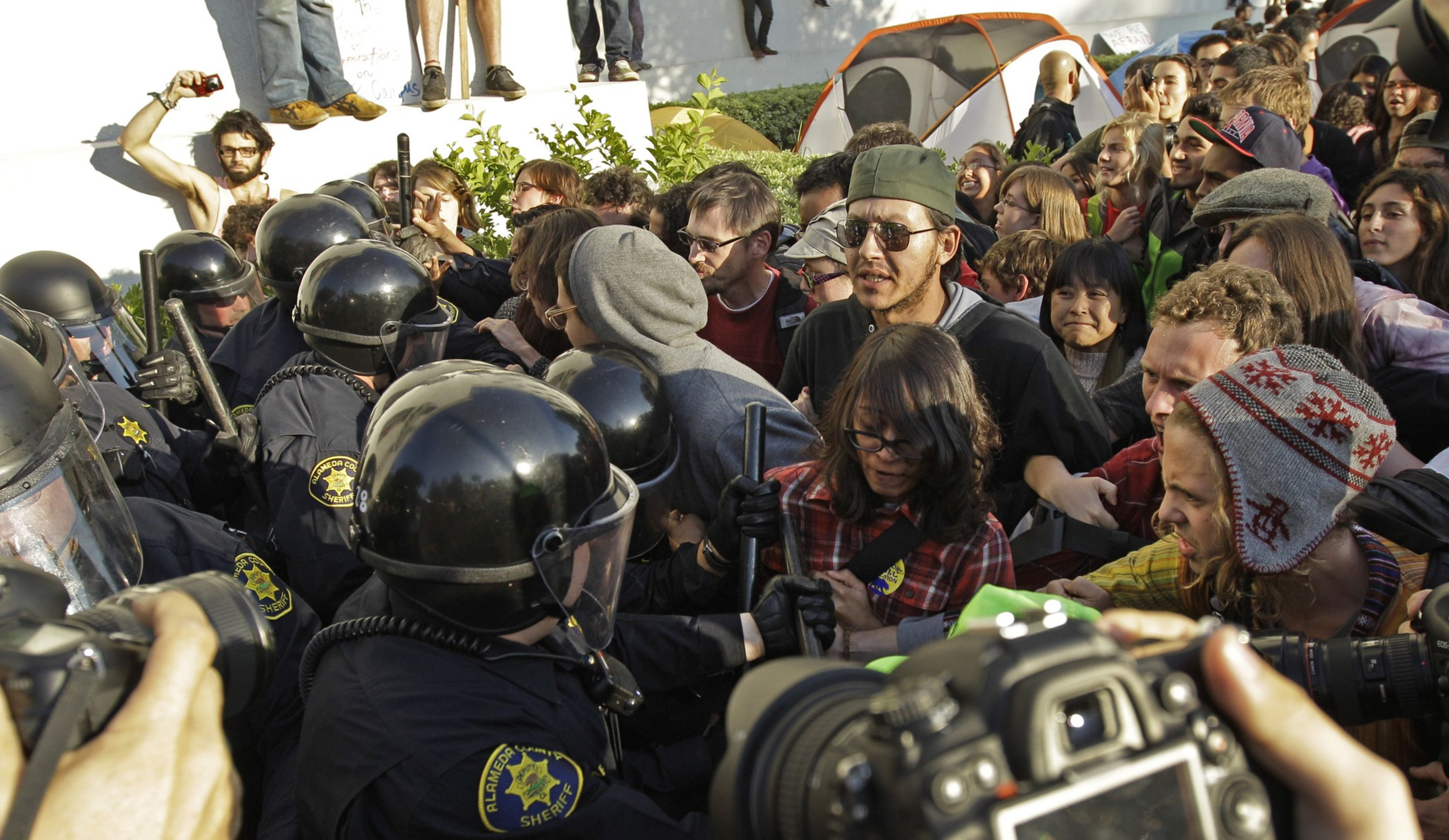A tense encounter between helmeted police and protesters with a background tense crowd and cameras.