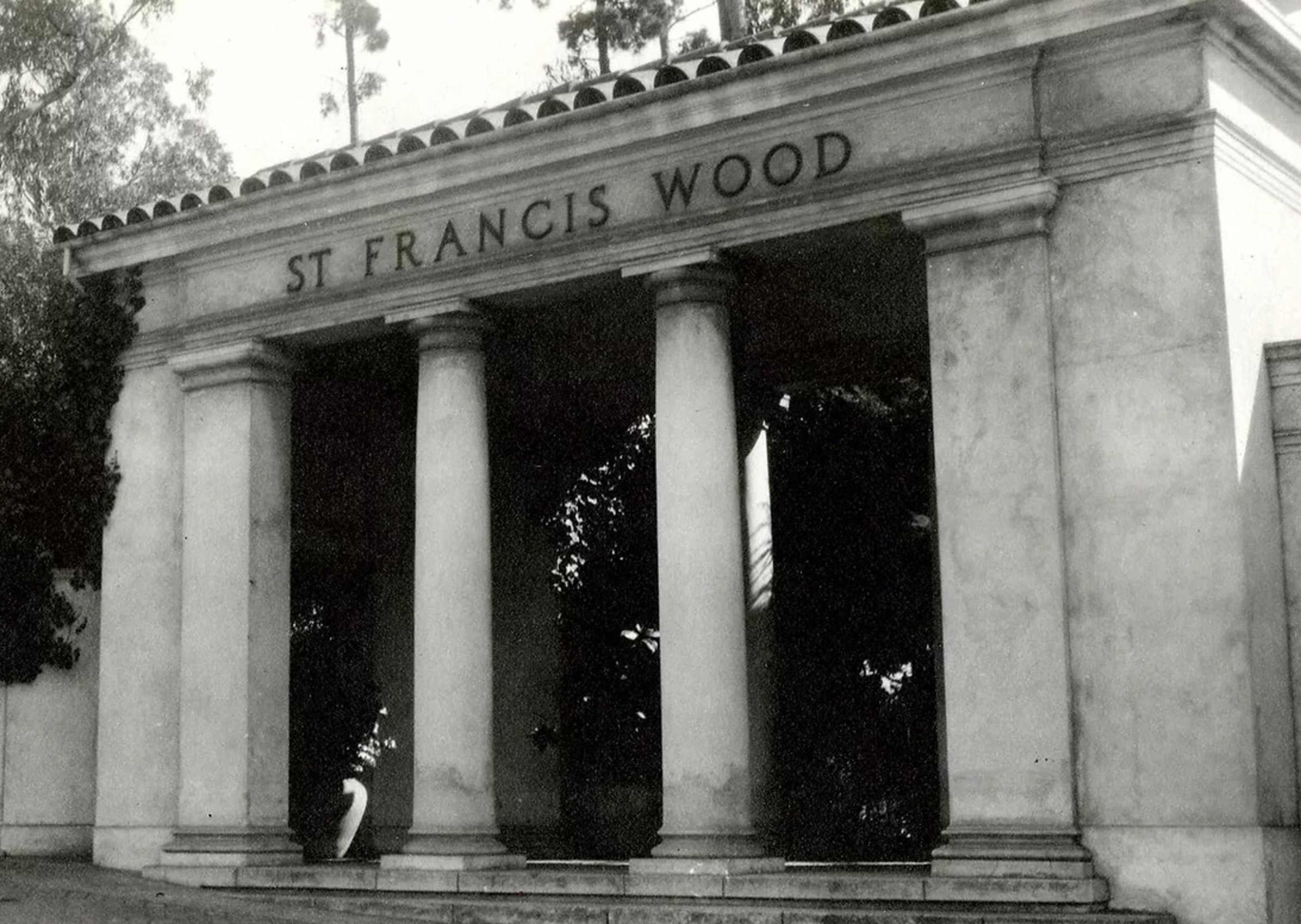 A stone entrance with four large pillars supports a roof labeled "ST FRANCIS WOOD," surrounded by trees and shadows.