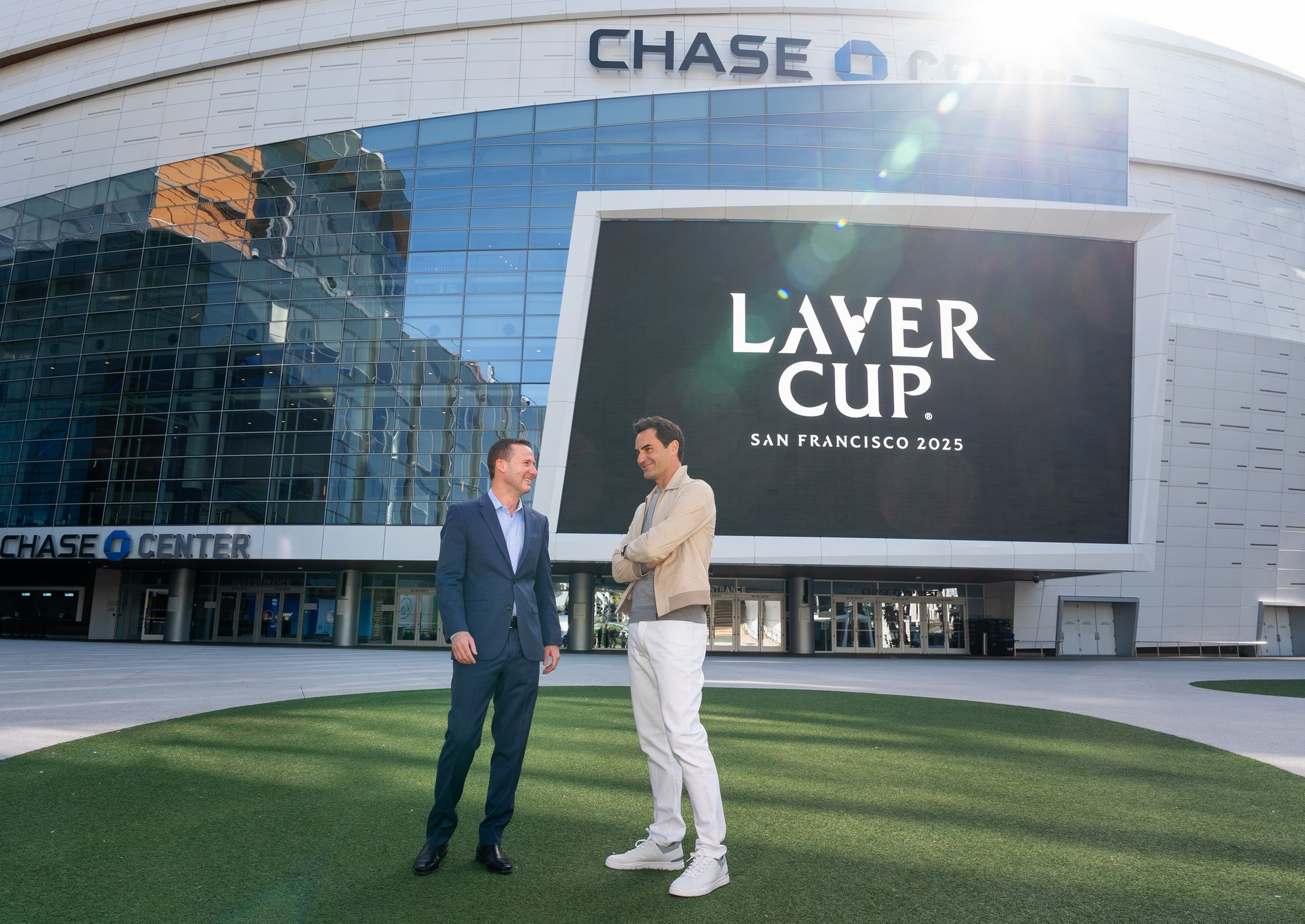 Two men stand smiling and talking on green grass outside Chase Center, with a large screen behind them showing “Laver Cup San Francisco 2025.”
