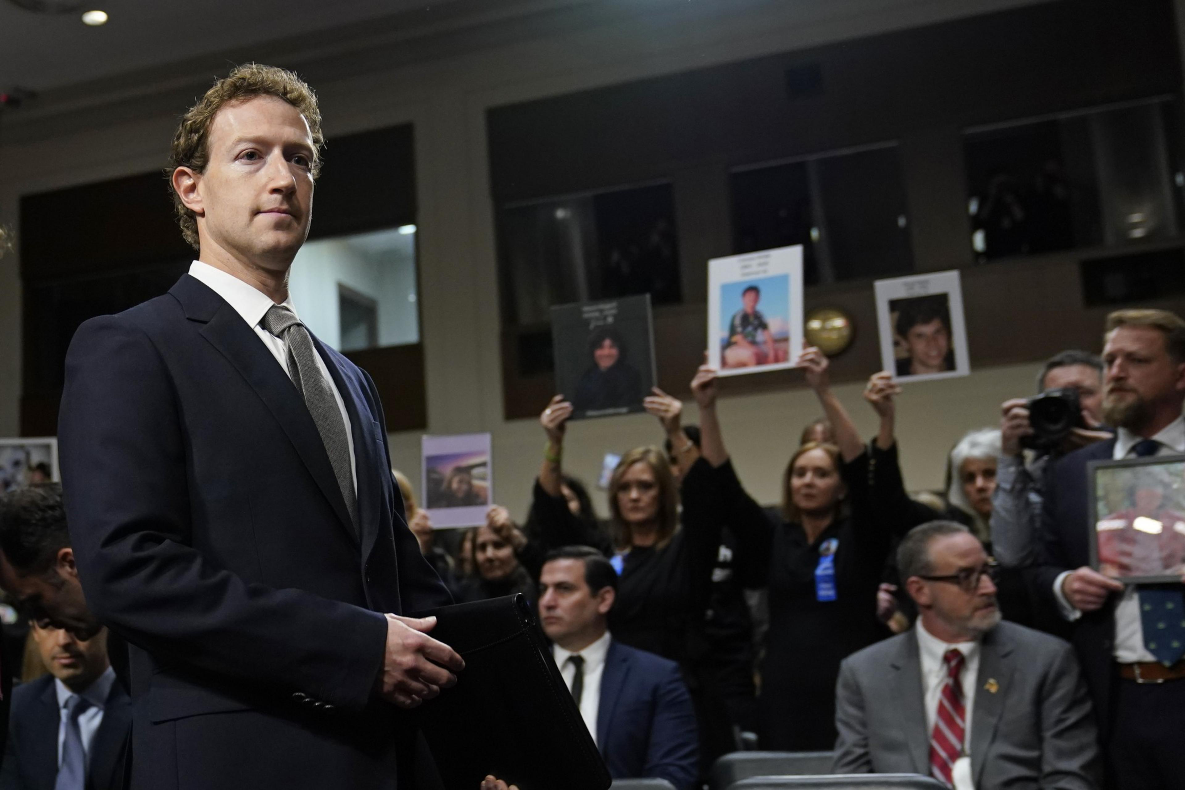 Man in suit and tie stands as women raise photos of young people in background