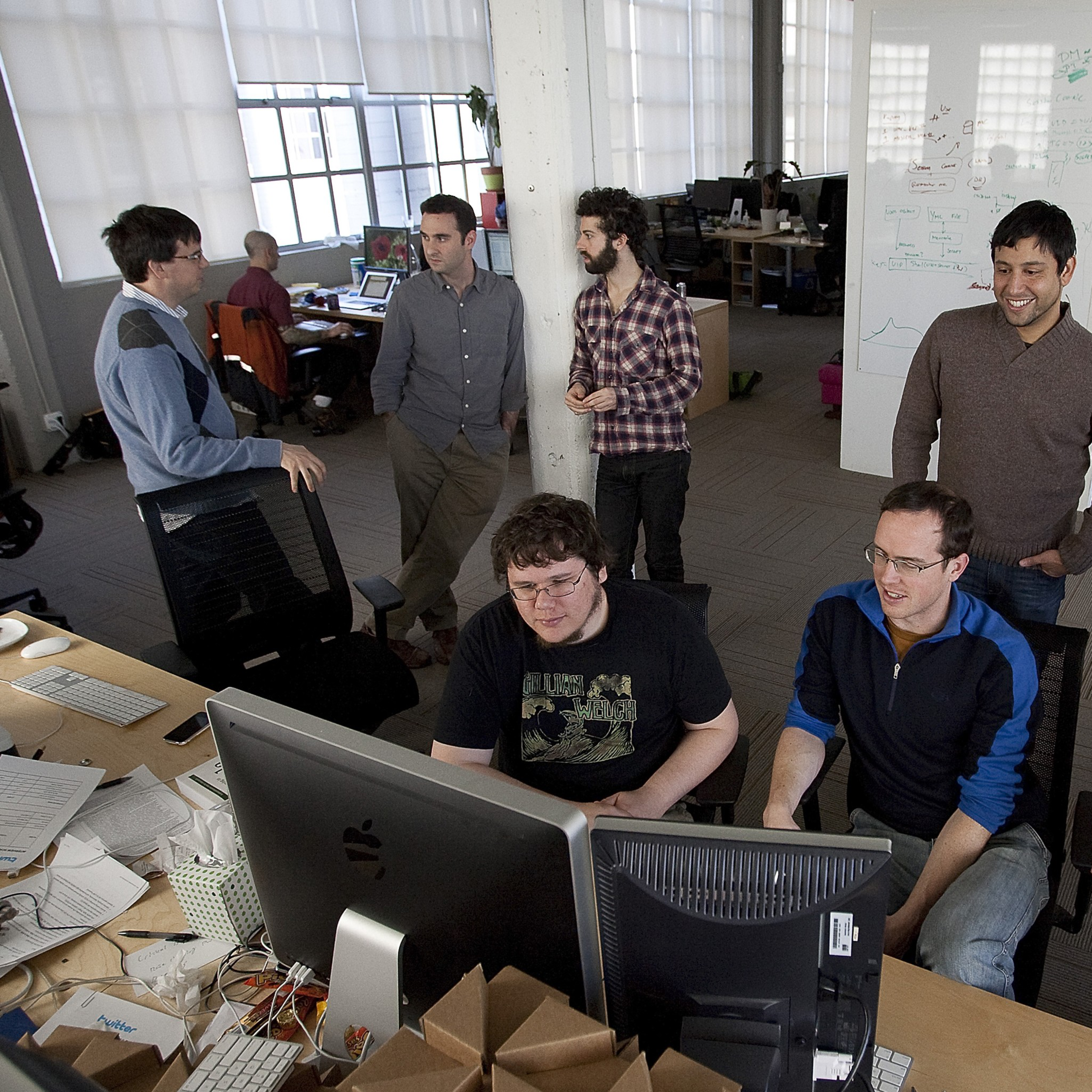 Six men in a modern office workspace are engaged in discussion and collaboration, with two sitting at computers and four standing nearby.