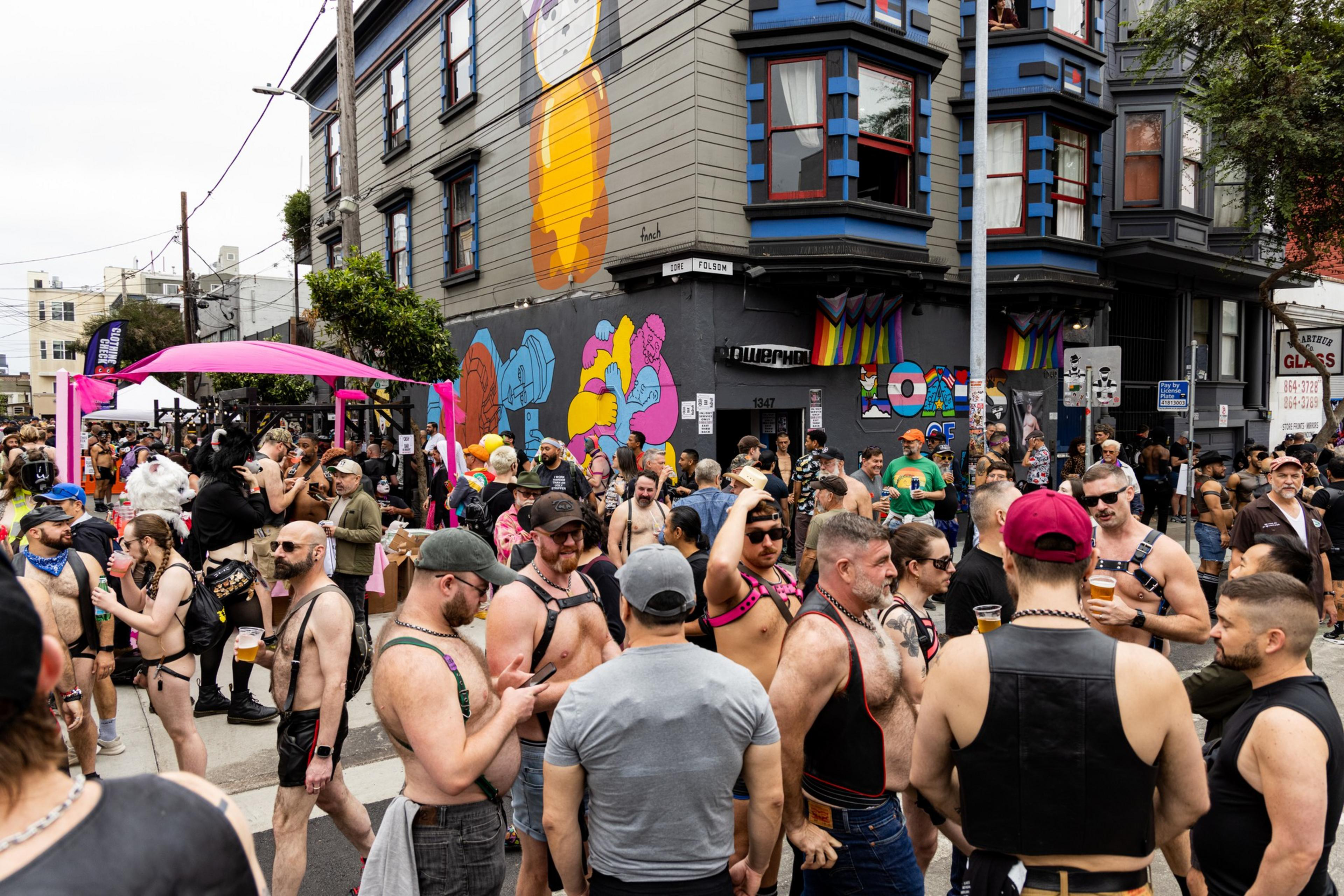 A lively street gathering with many shirtless men wearing leather harnesses and casual clothing, colorful murals on the building, and a pink canopy in the background.
