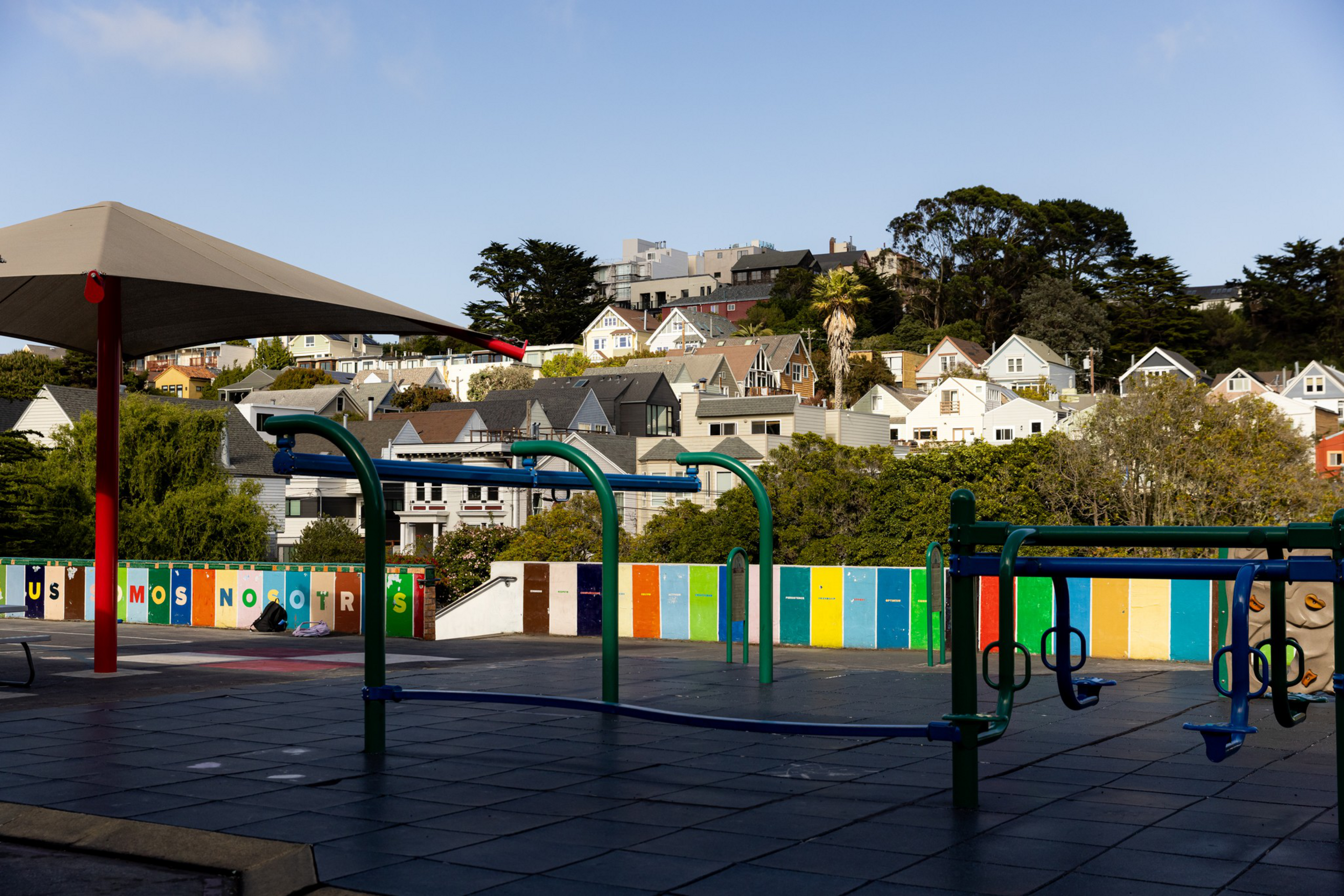 A colorful playground with climbing structures and swings sits empty in front of a row of residential houses under a clear sky.