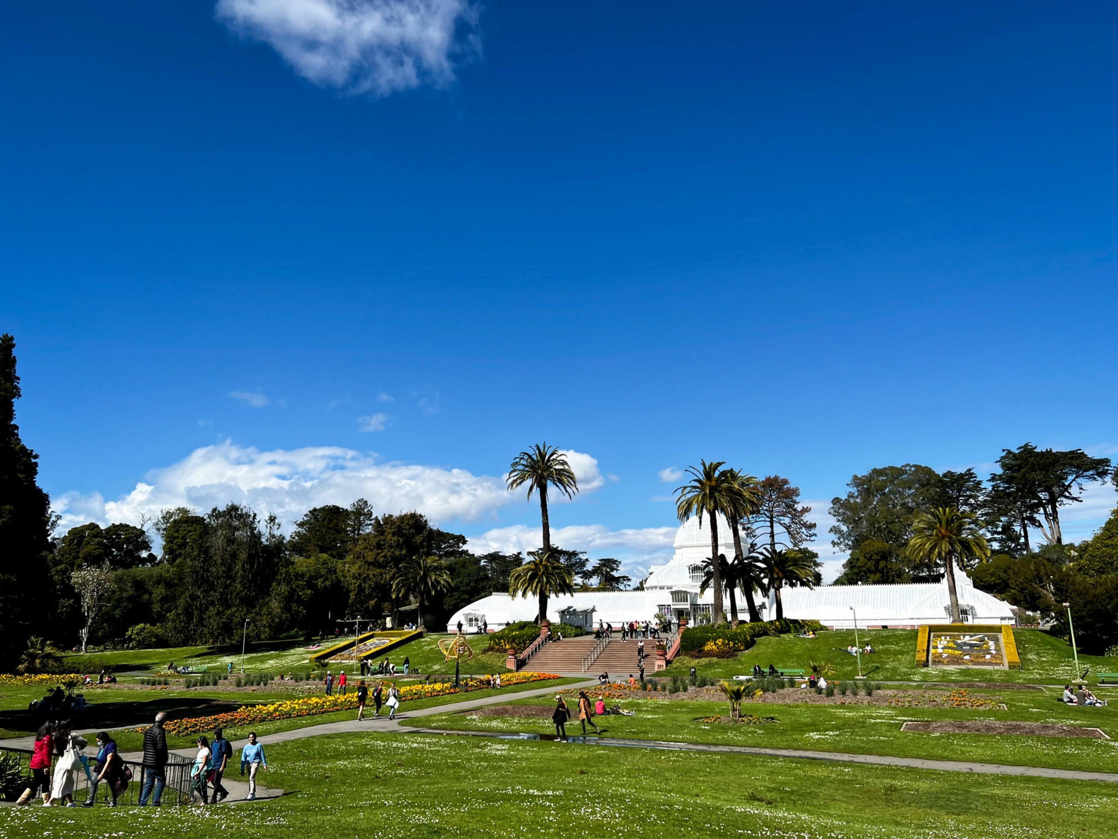 A white greenhouse with a dome sits at the center of a park with palm trees, flower beds, and people walking or sitting on grassy lawns under a clear blue sky.