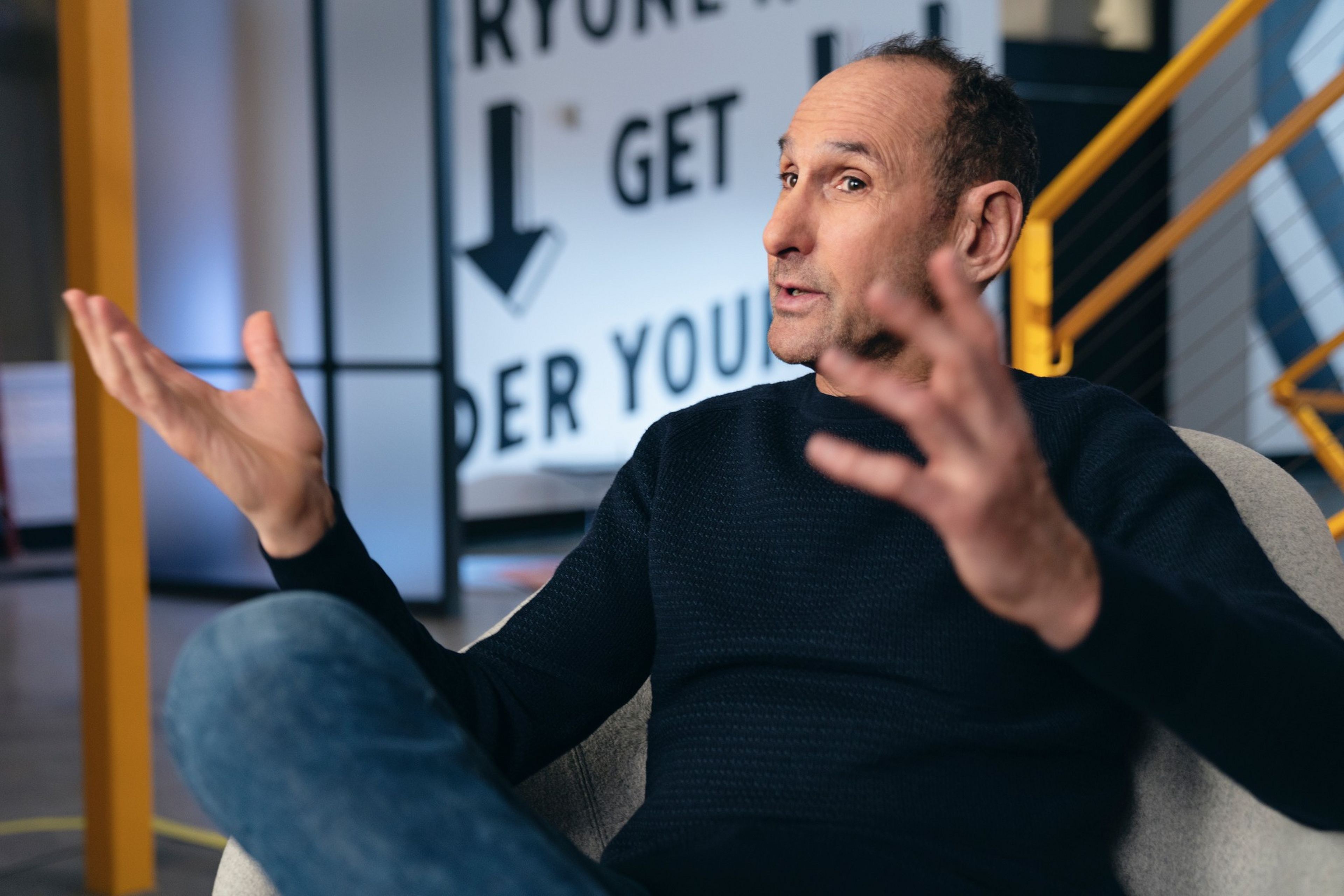 A man in a dark sweater and jeans sits in a chair, gesturing with both hands, with a blurred sign and yellow railing in the background.