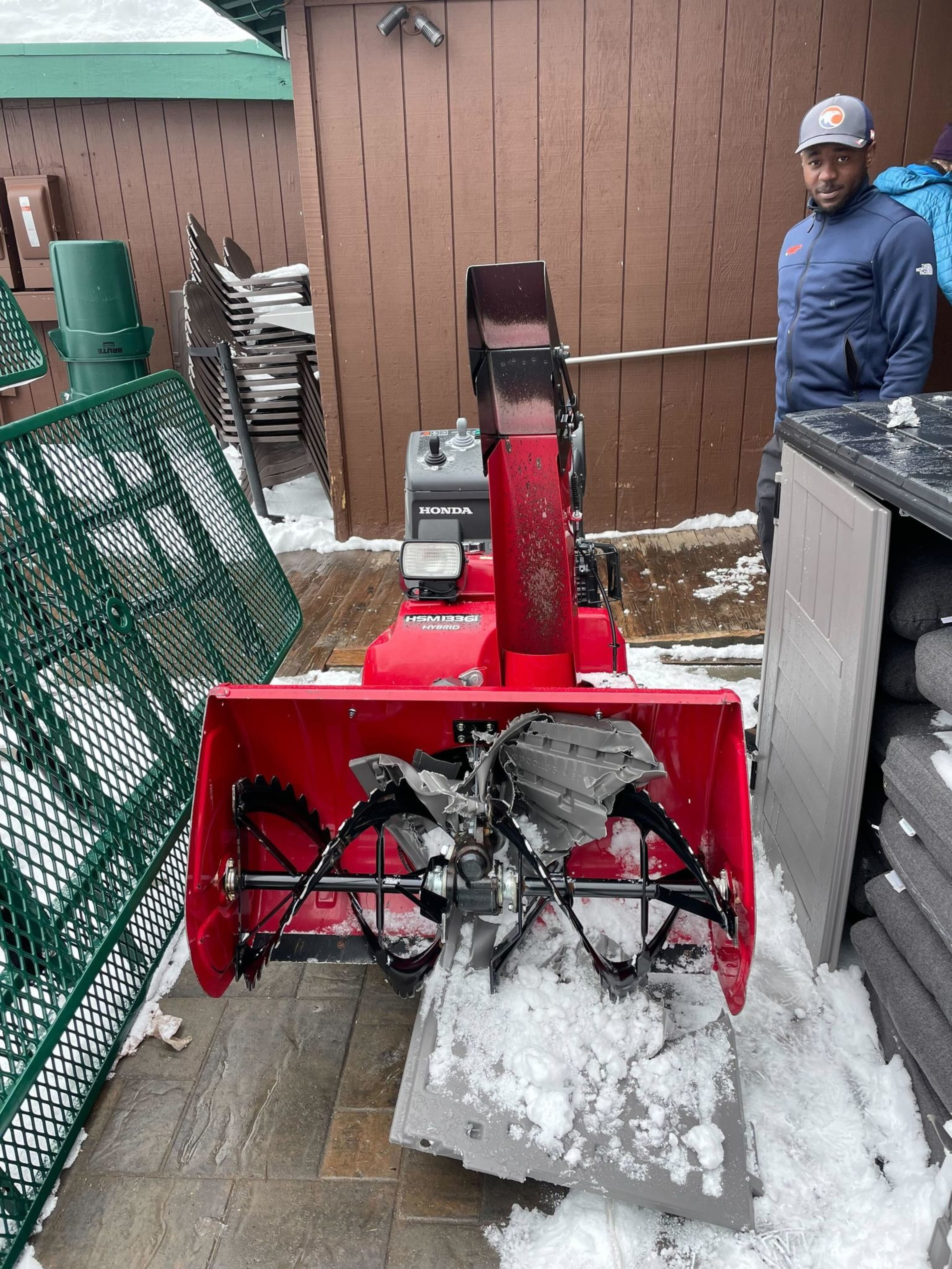 A red snow blower stands on a snowy patio, surrounded by brown fencing and green metal railings. A person in a blue jacket and cap is nearby.