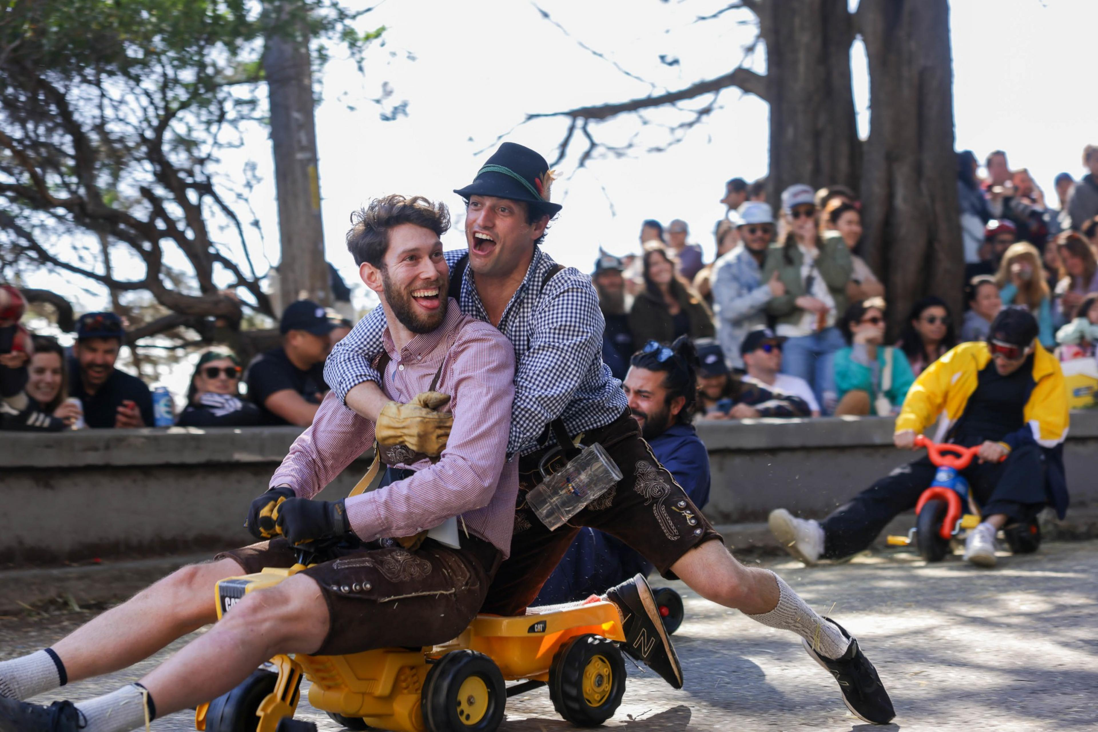 Two men ride a small yellow plastic toy truck, smiling joyously, as spectators watch in the background.
