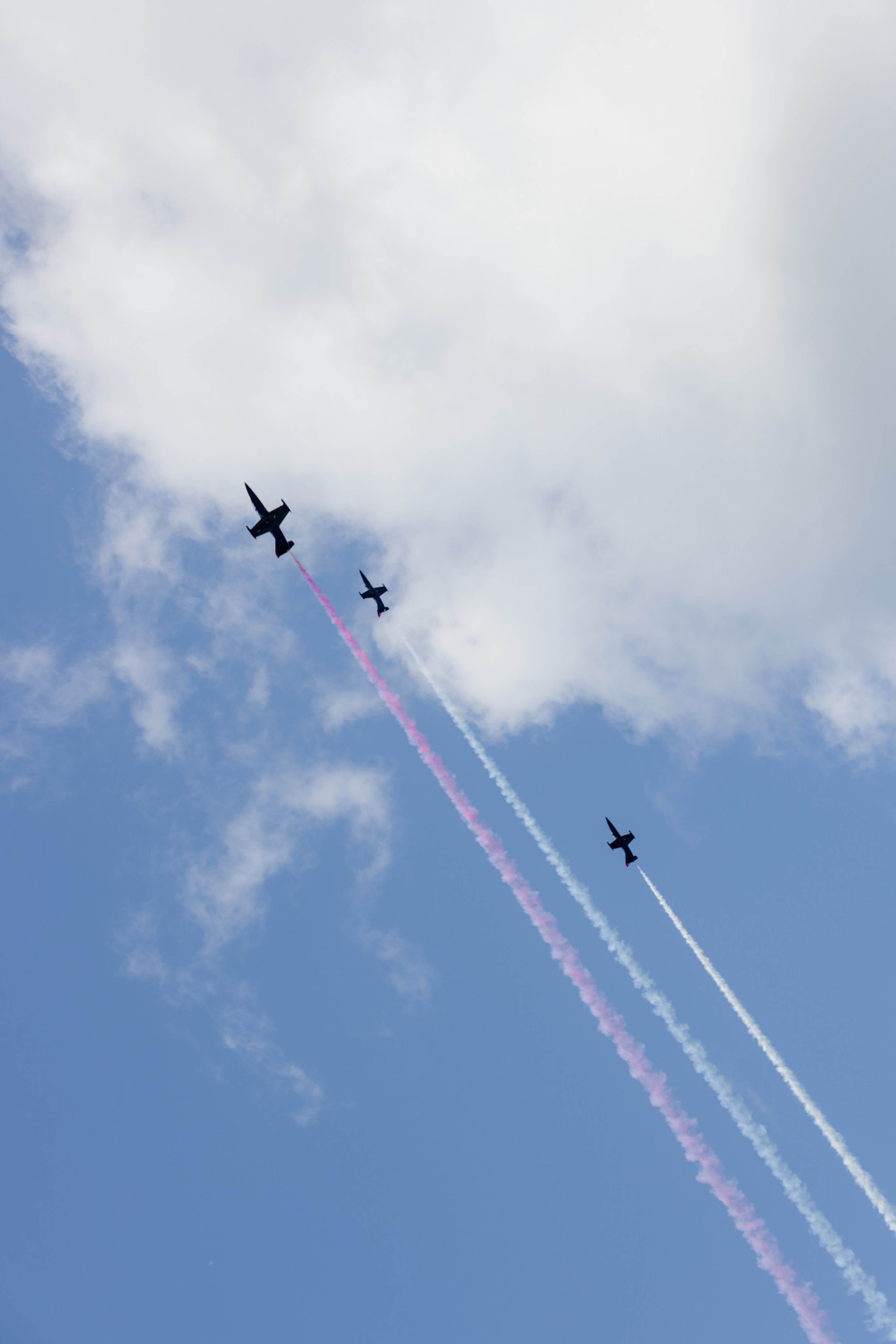 Three jets fly upward in a clear blue sky with some clouds, leaving behind trails of pink, white, and blue smoke.