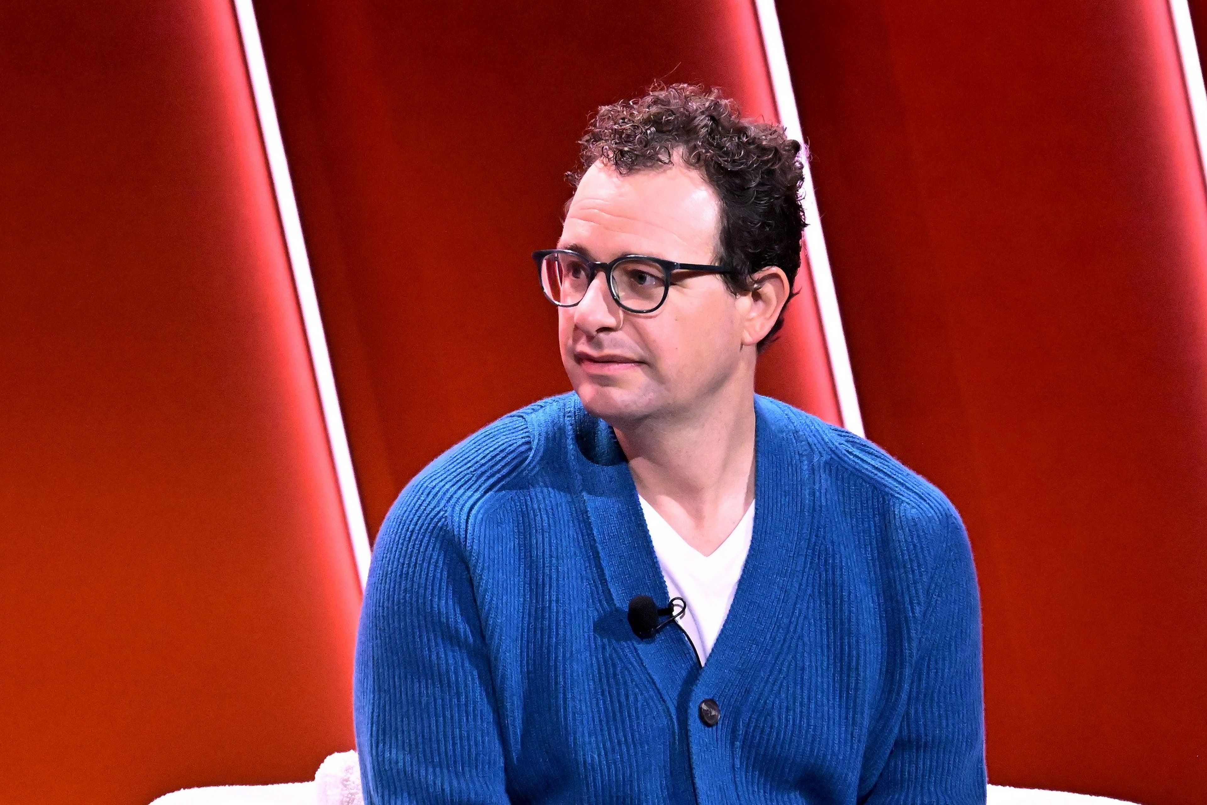 A man with curly dark hair and glasses wears a blue cardigan over a white shirt, sitting against a red background with vertical light strips.