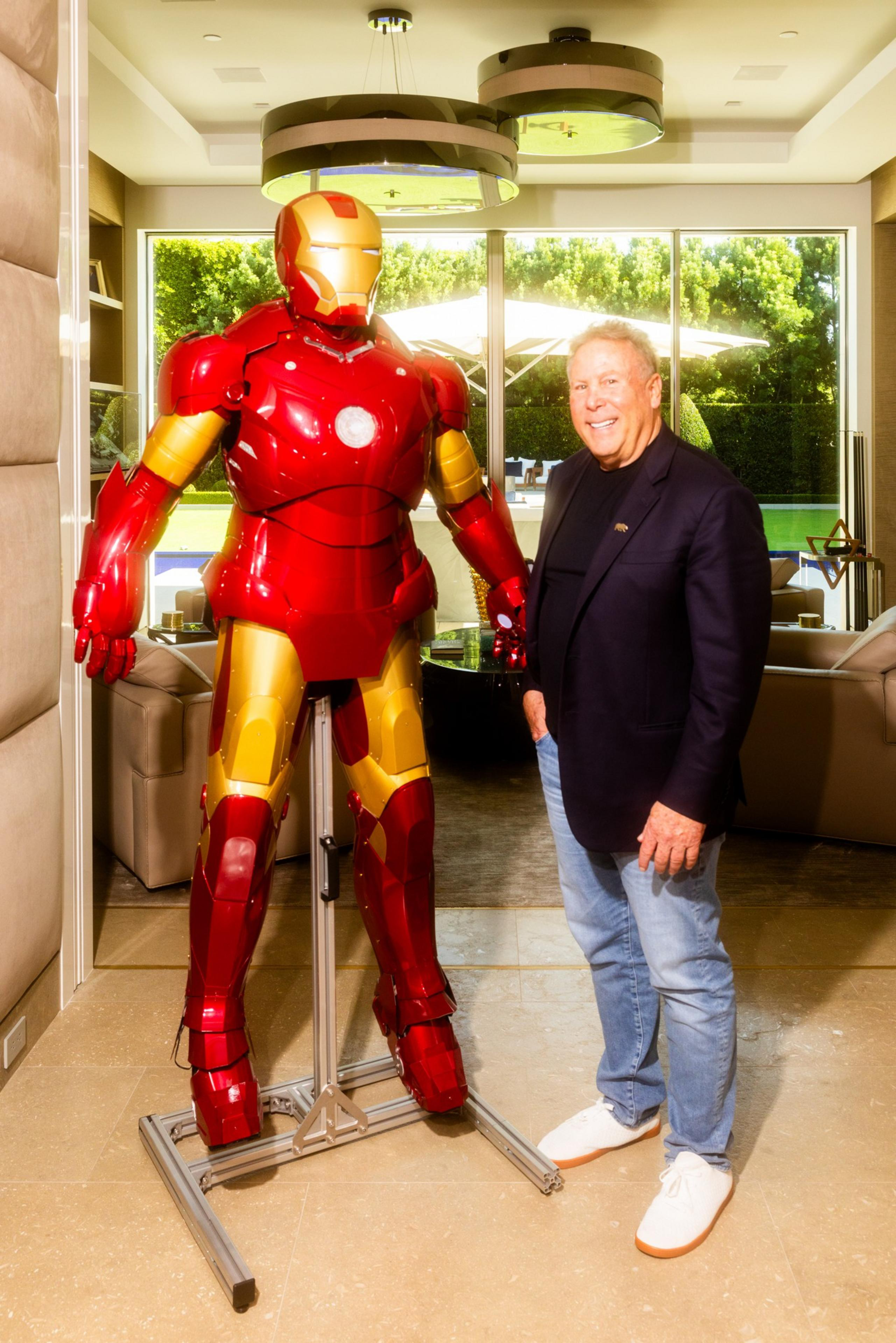 A man in a blazer and jeans stands smiling beside a life-sized Iron Man statue inside a sunlit, modern living room.