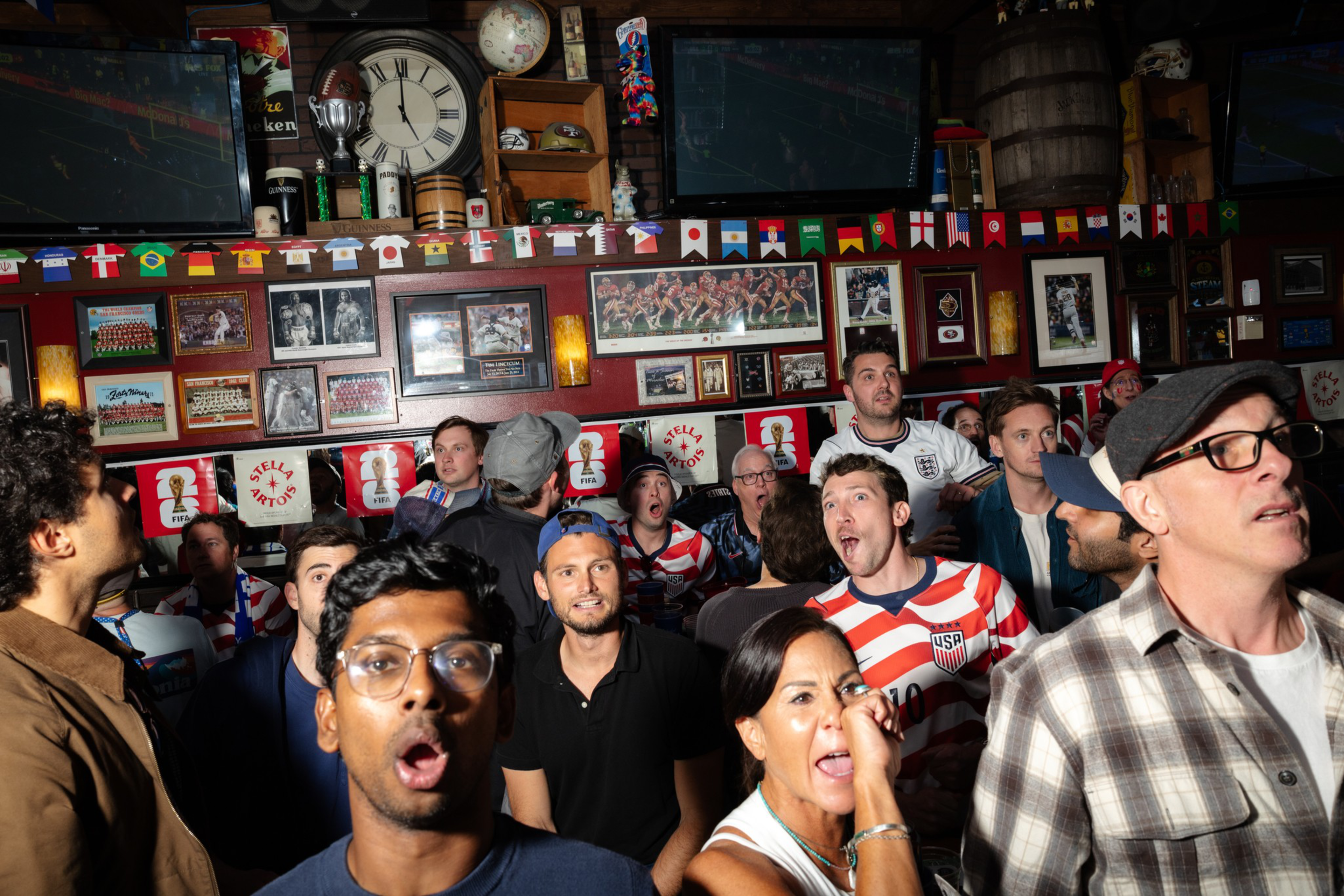 A crowded bar with diverse people watching a sports game intensely, some wearing soccer jerseys, surrounded by sports memorabilia and flags.