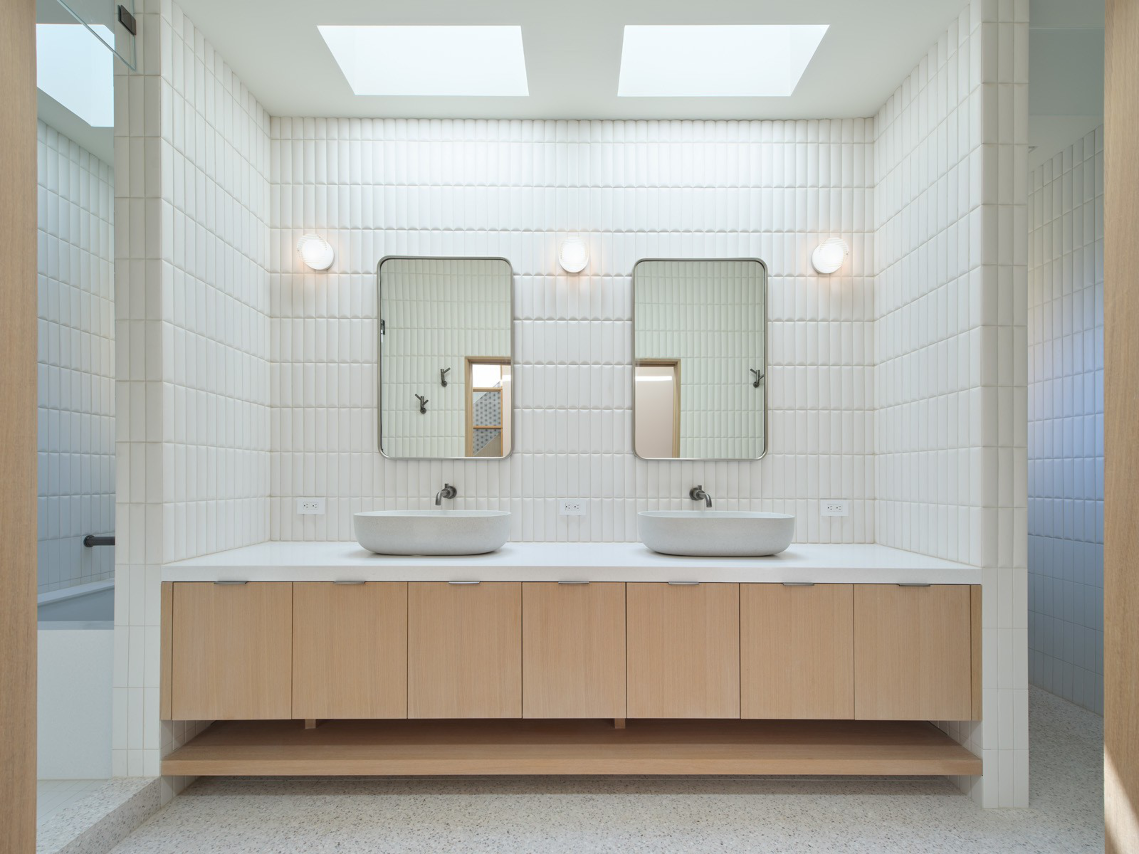 A minimalist bathroom features two countertop vessel sinks, rectangular mirrors with rounded edges, wooden cabinets below, white vertically tiled walls, and skylights above.