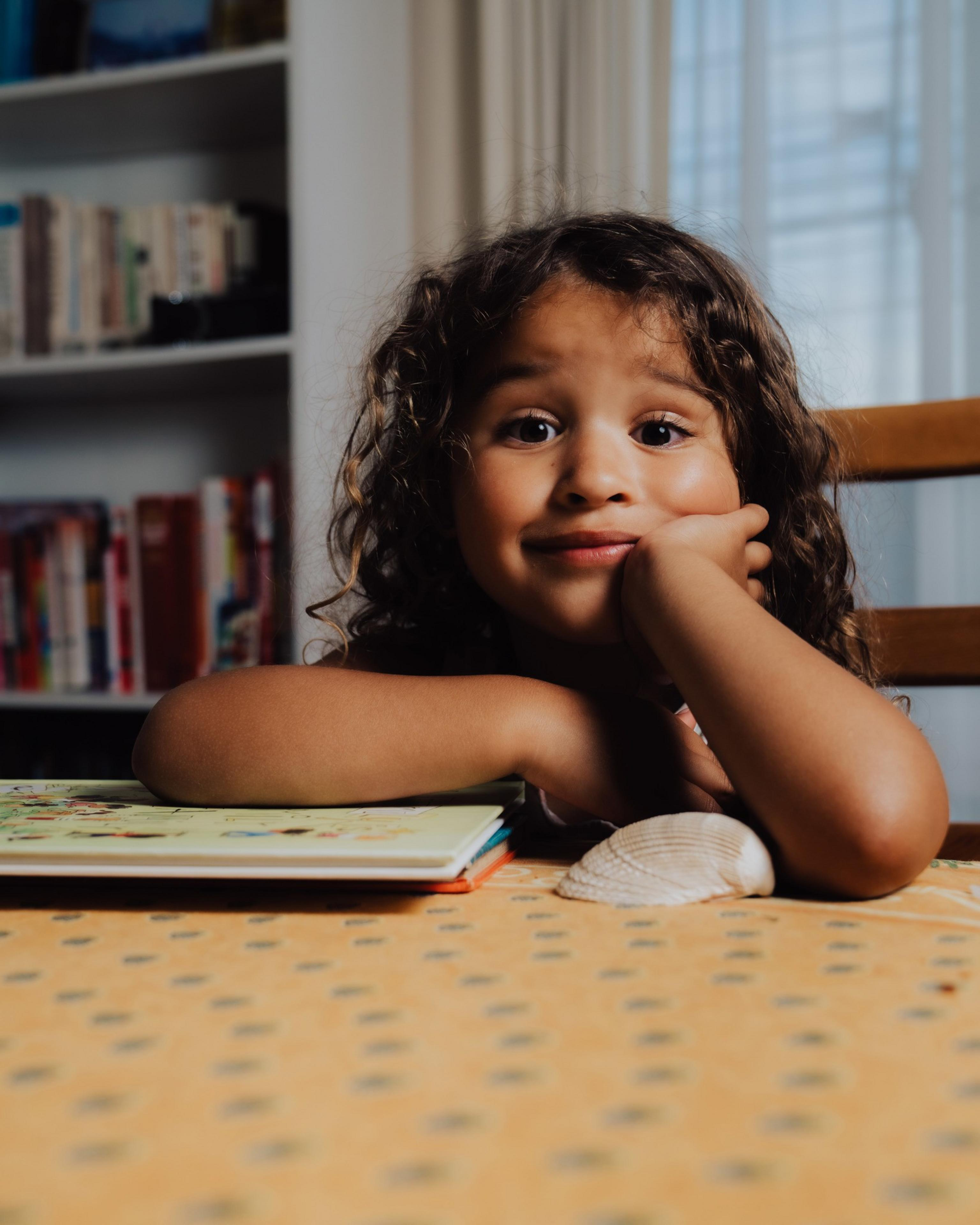 A child with curly hair rests their chin on a hand, sitting at a table with a book and a seashell. Shelves filled with books are visible in the background.