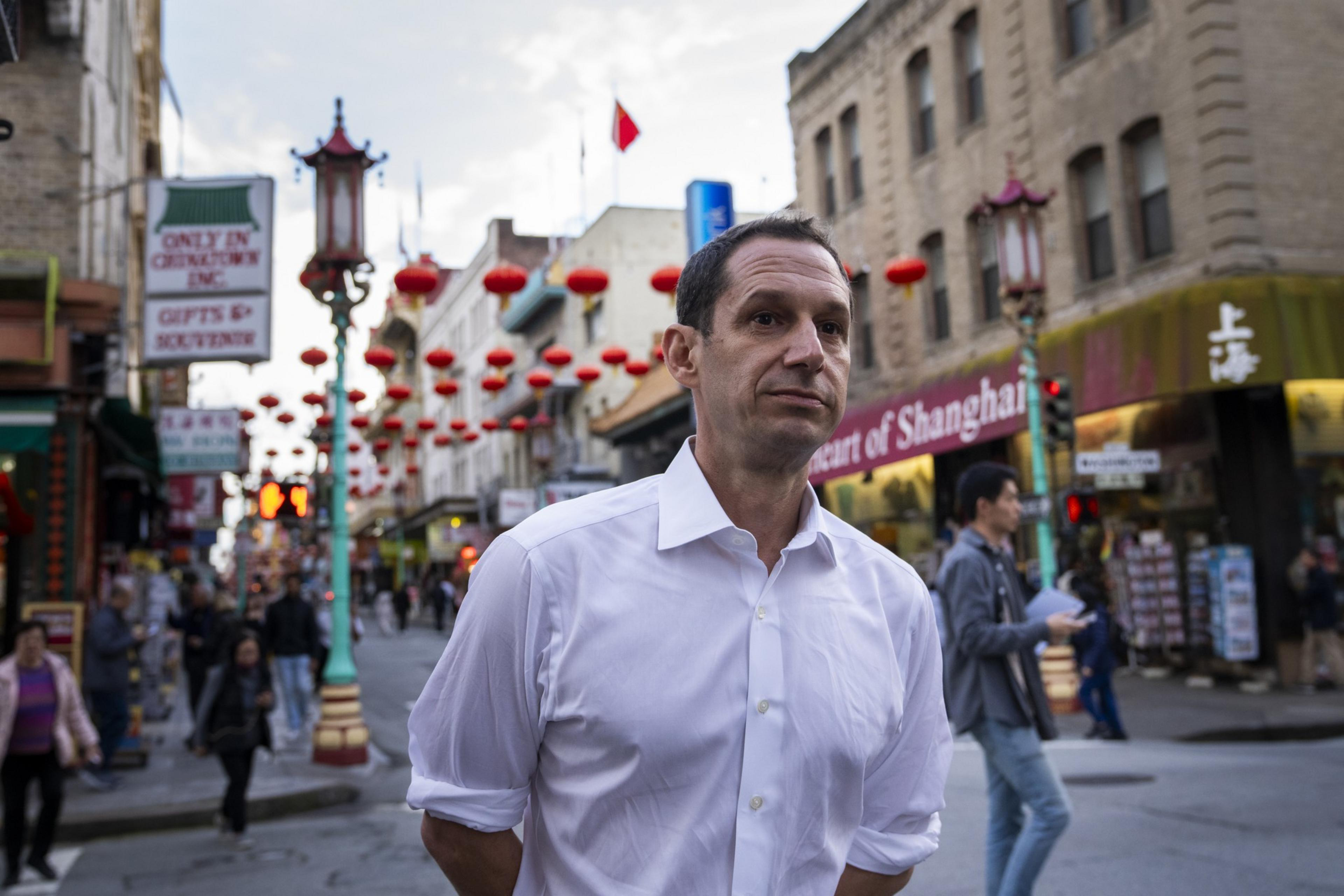 A man in a white shirt stands on a bustling street decorated with red lanterns, likely in a Chinatown. People walk by, and signs advertise gifts and Shanghai.