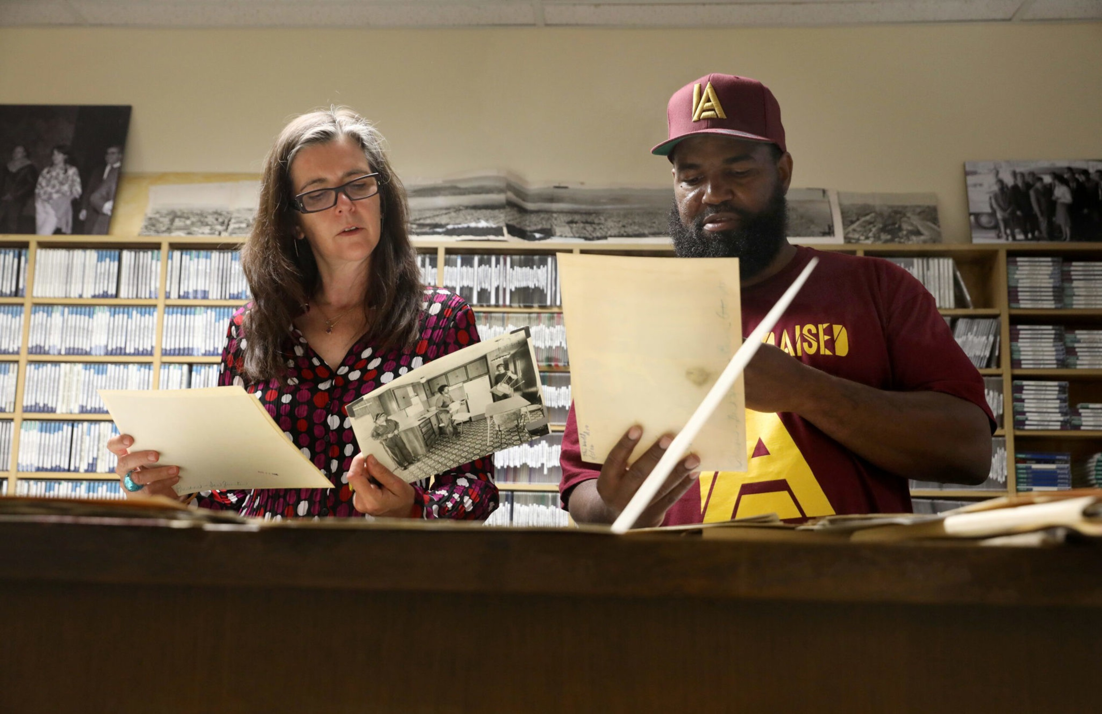 Two people are examining old photographs and documents in a room with shelves of CDs and an image of musicians behind them.