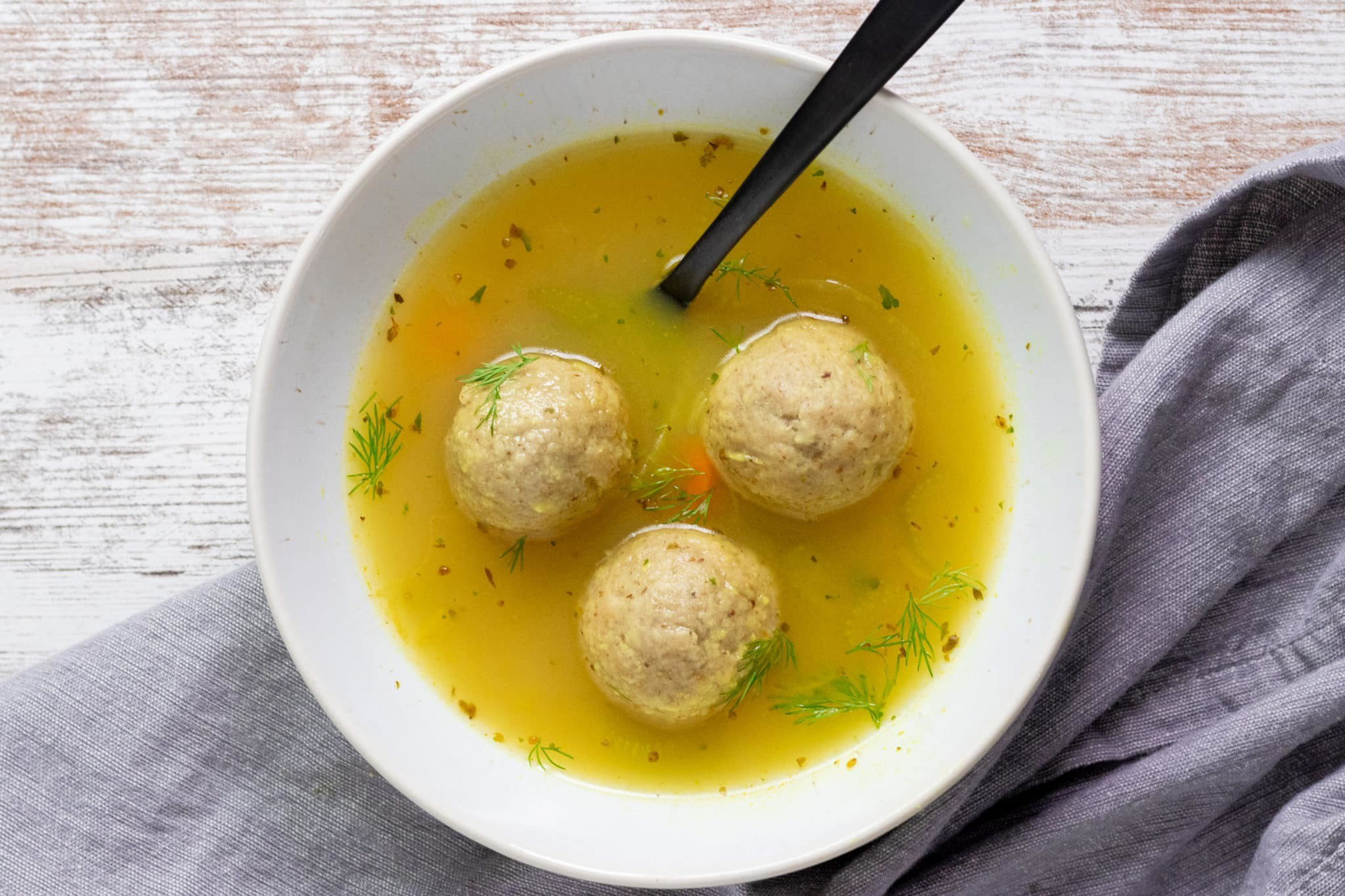 A white bowl holds three matzo balls in clear broth with dill and carrot pieces, a black spoon resting inside, set on a gray cloth on a wooden surface.