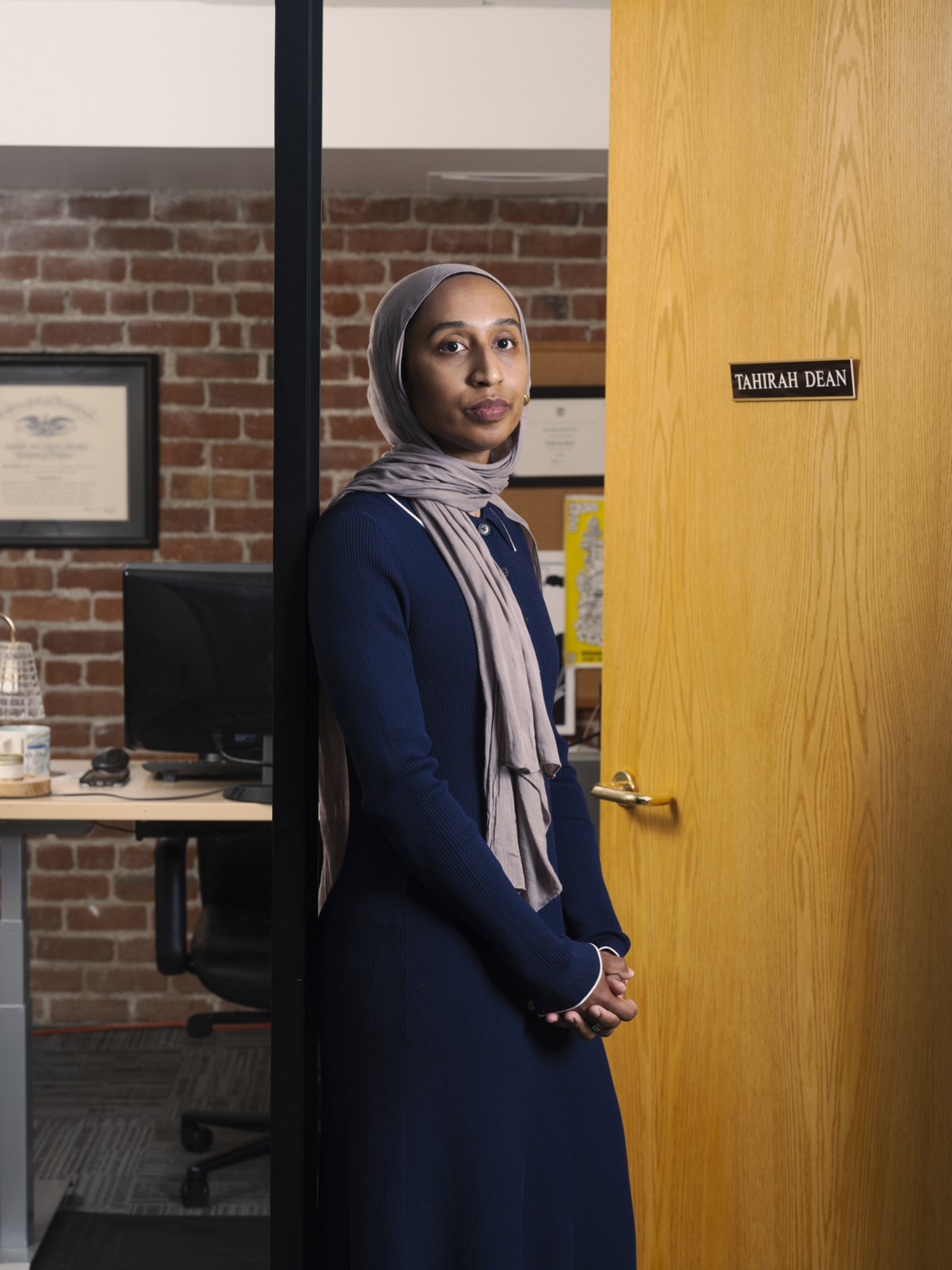 A person in a blue outfit and beige headscarf stands by an office door labeled &quot;Tahirah Dean.&quot; Behind them is a brick wall and a desk with a computer.