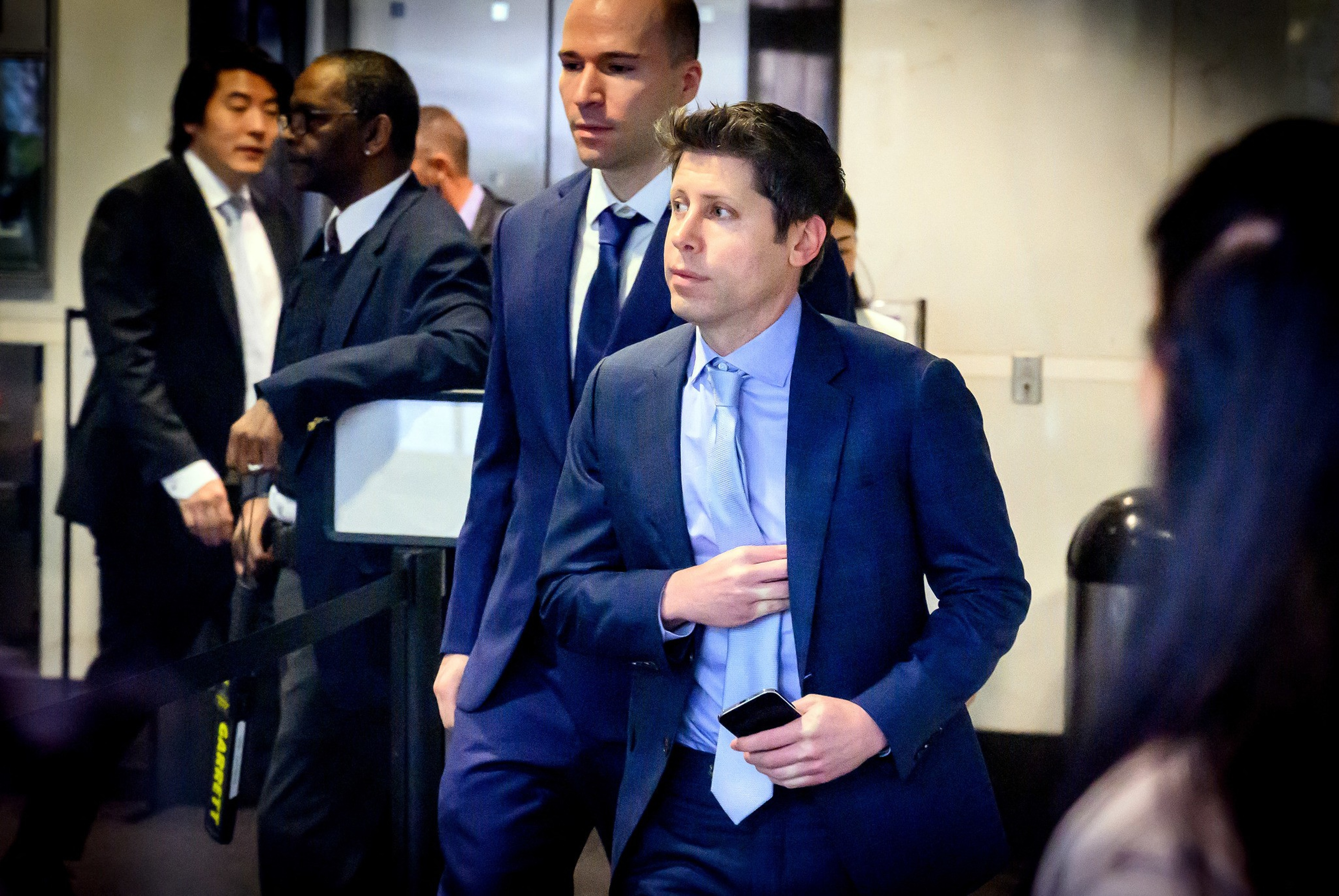 Several men in suits stand and walk indoors, one holding a smartphone and adjusting his jacket, while others wait or move in the background.