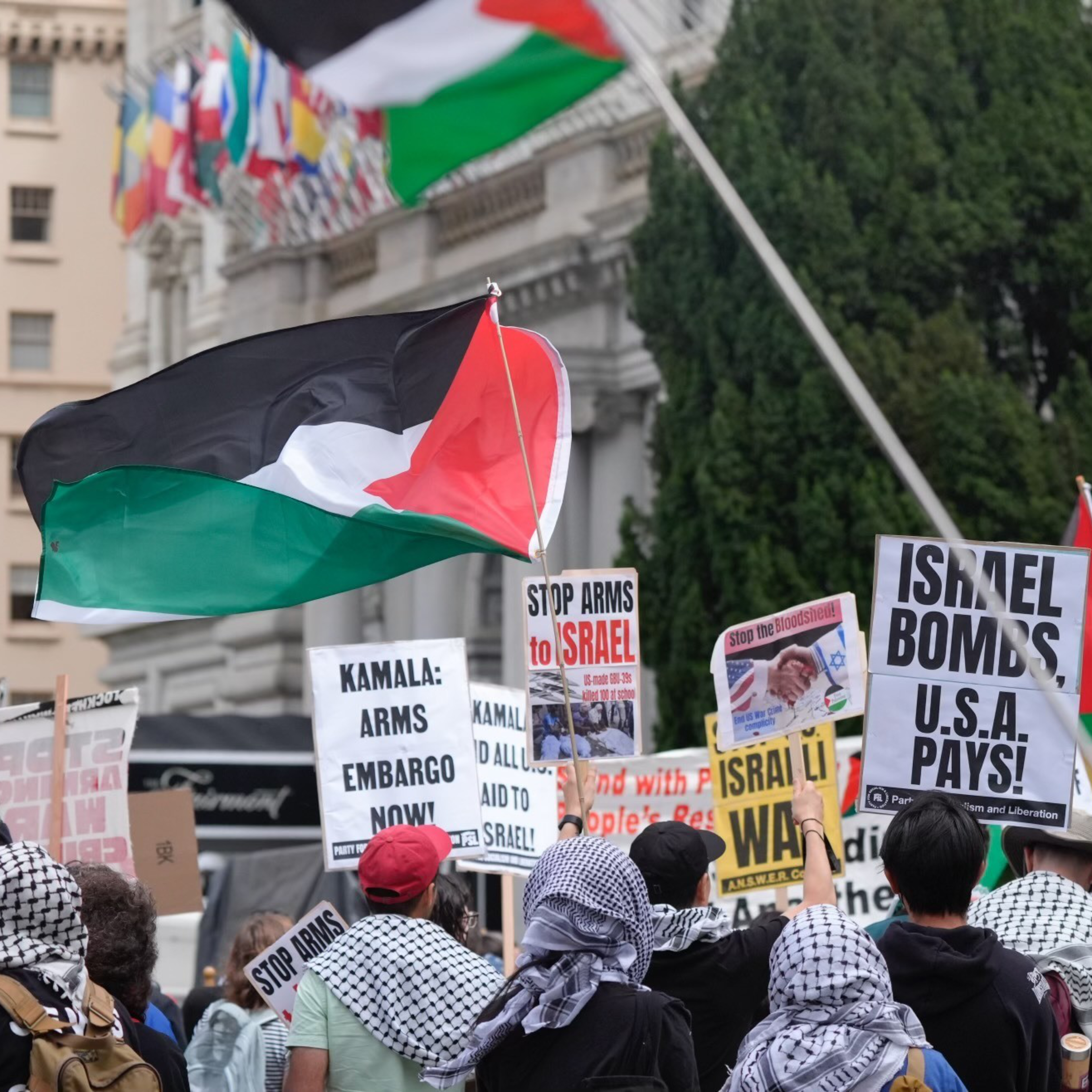 Protesters holding Palestinian flags and signs such as &quot;Stop Arms to Israel&quot; and &quot;Israel Bombs, U.S.A Pays!&quot; gather near a building and intersection named &quot;California&quot;.