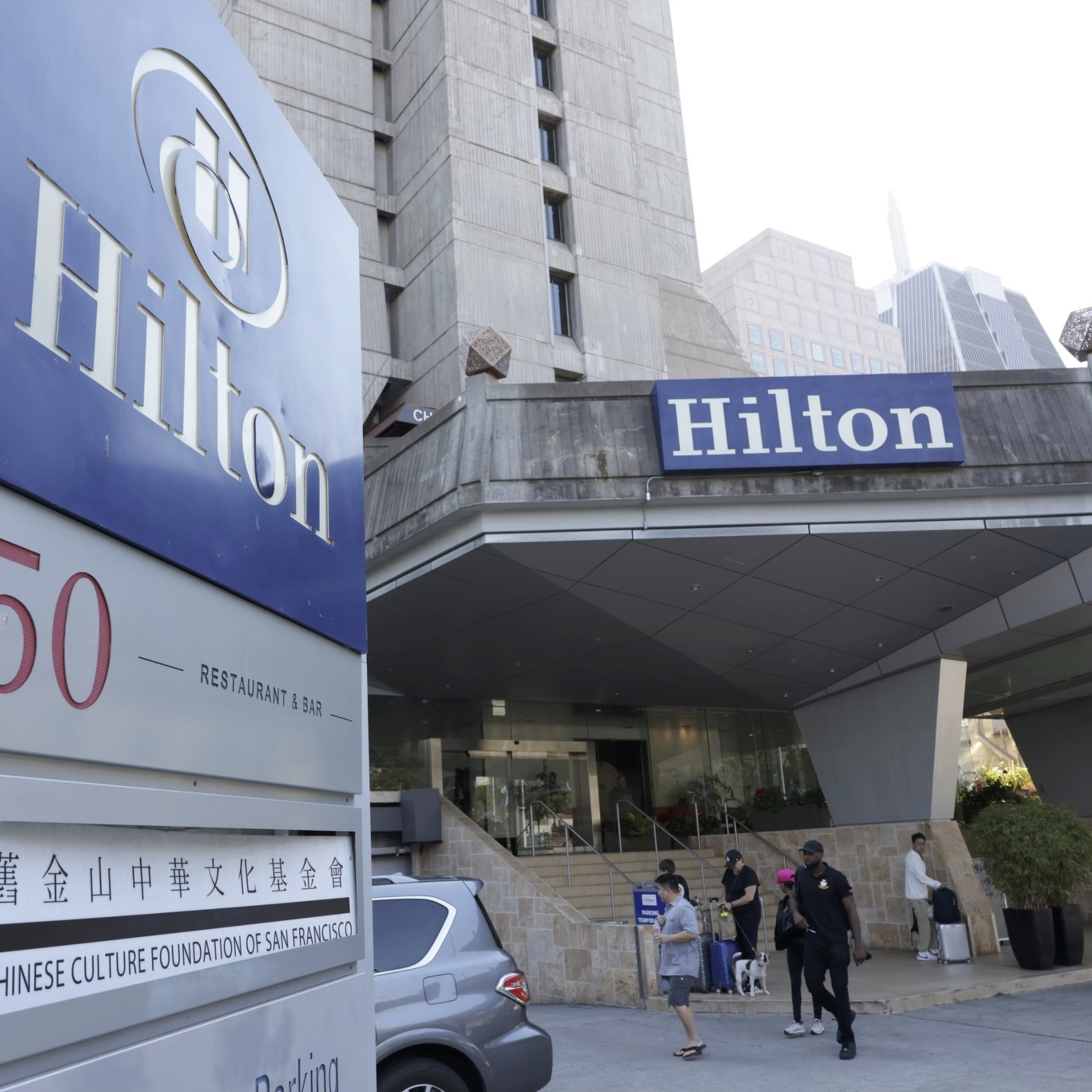 This image shows the entrance of a Hilton hotel with a large blue Hilton sign and the address number 750. People are walking and carrying luggage outside. The building is tall and modern.