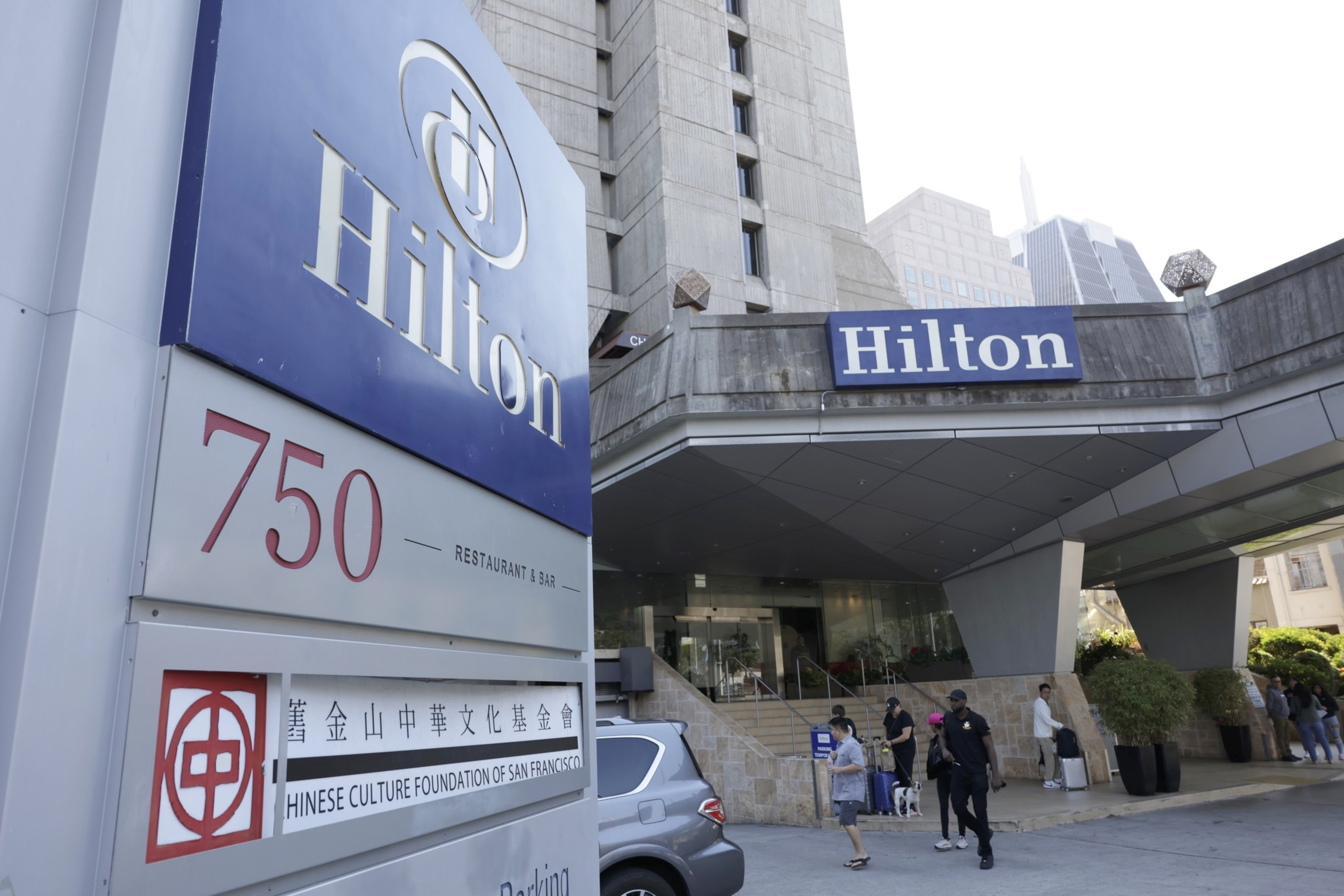 This image shows the entrance of a Hilton hotel with a large blue Hilton sign and the address number 750. People are walking and carrying luggage outside. The building is tall and modern.