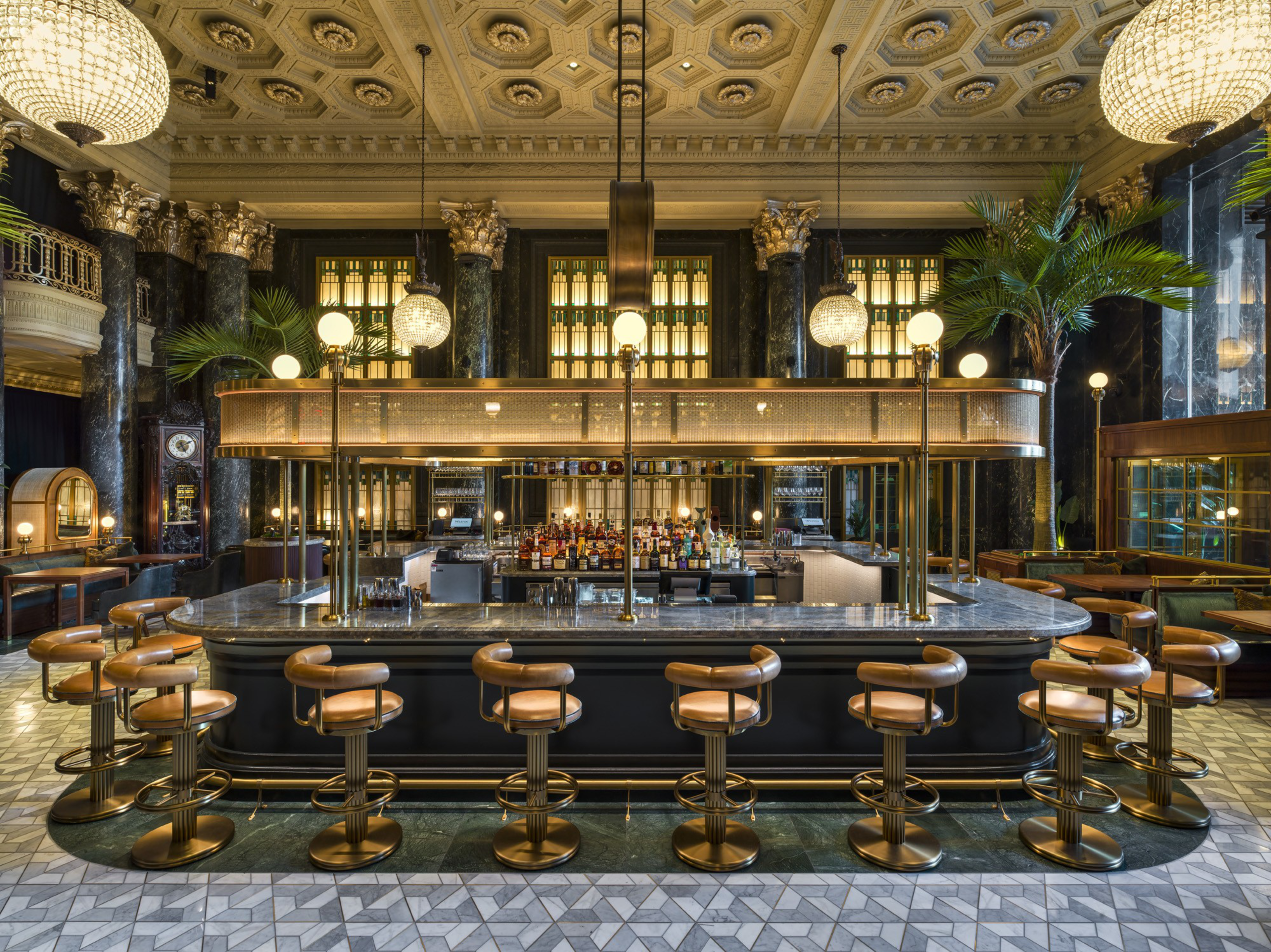 A luxurious bar with a marble counter, surrounded by tan leather stools, ornate columns, globe lights, and a richly decorated coffered ceiling.