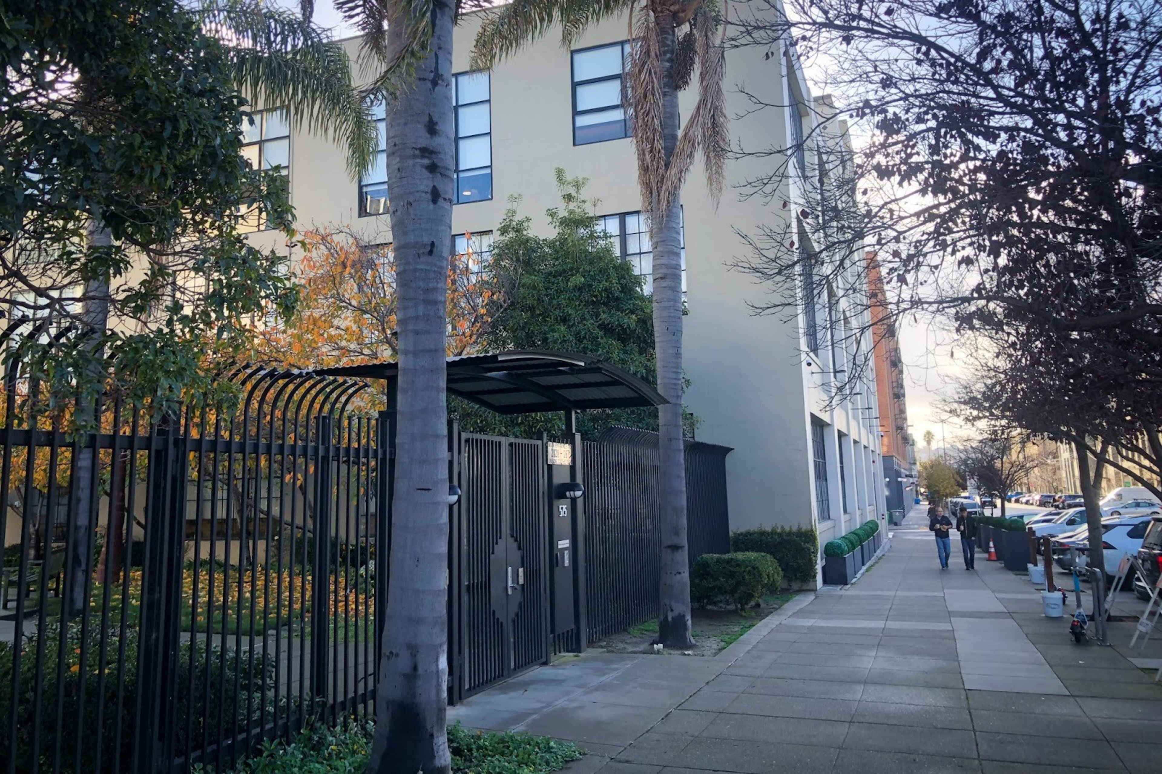 A gated entrance with a curved black metal awning stands between two tall palm trees along a sidewalk next to a multi-story building.