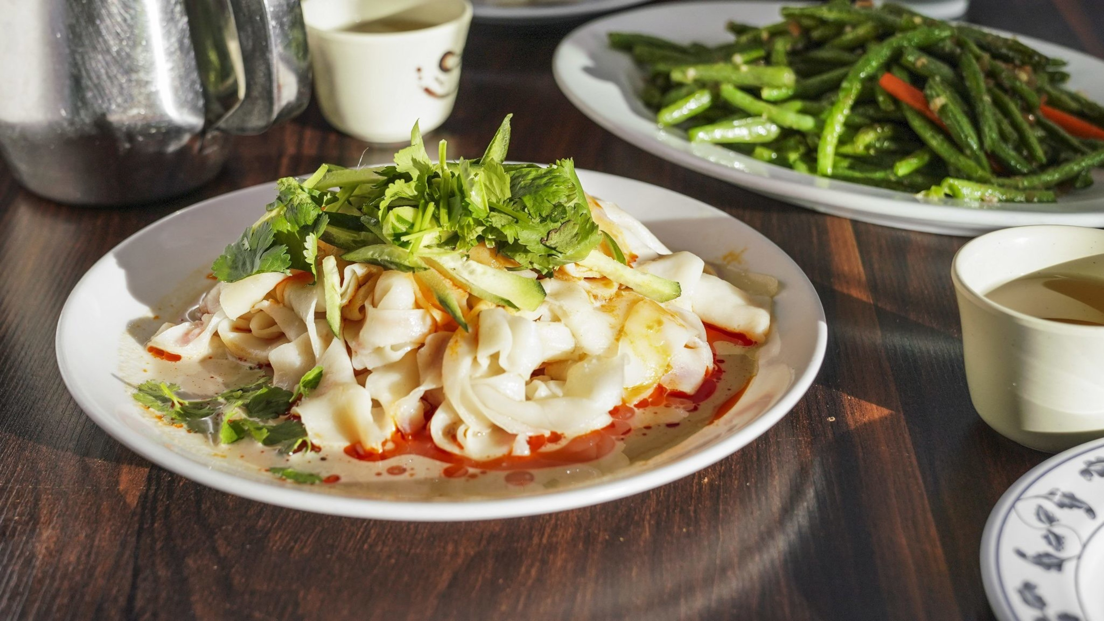 The image shows a plate of wide noodles topped with cilantro and cucumber, drizzled with chili oil. In the background, there’s a dish of stir-fried green beans.
