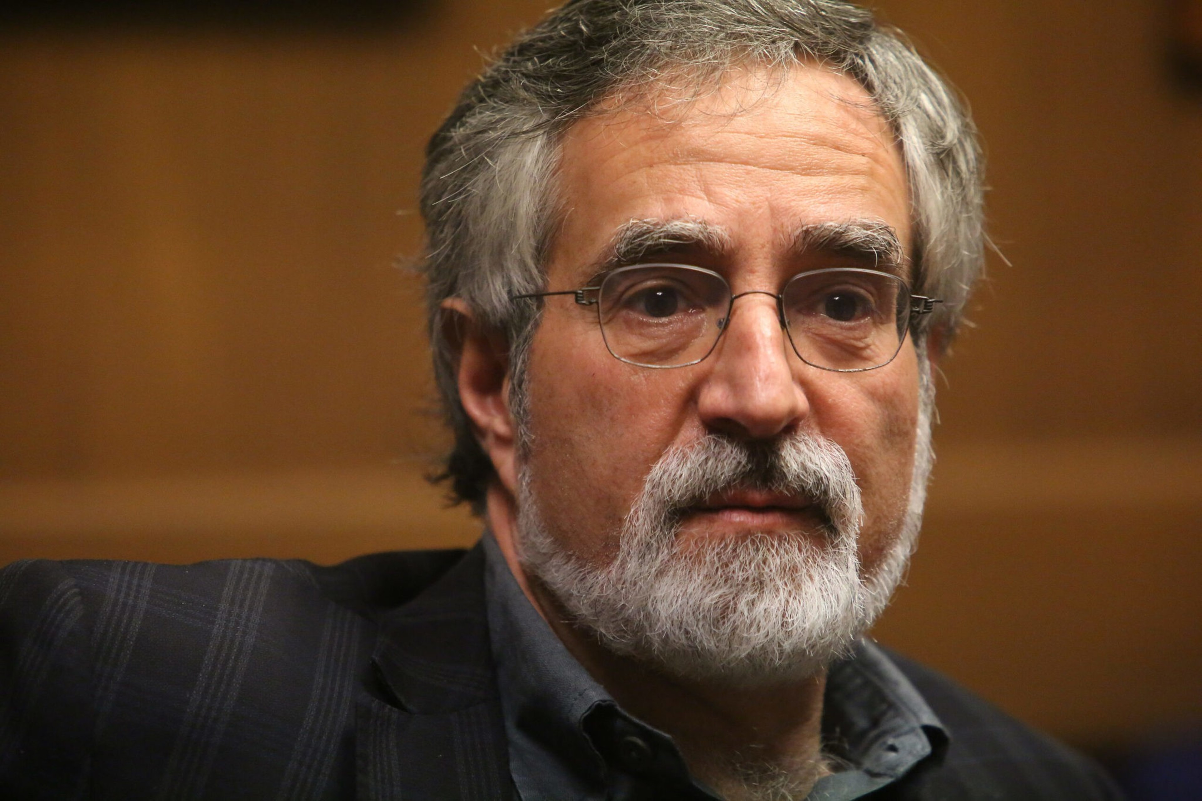 A bearded, bespectacled man with salt-and-pepper hair, wearing a dark suit, is captured mid-conversation.