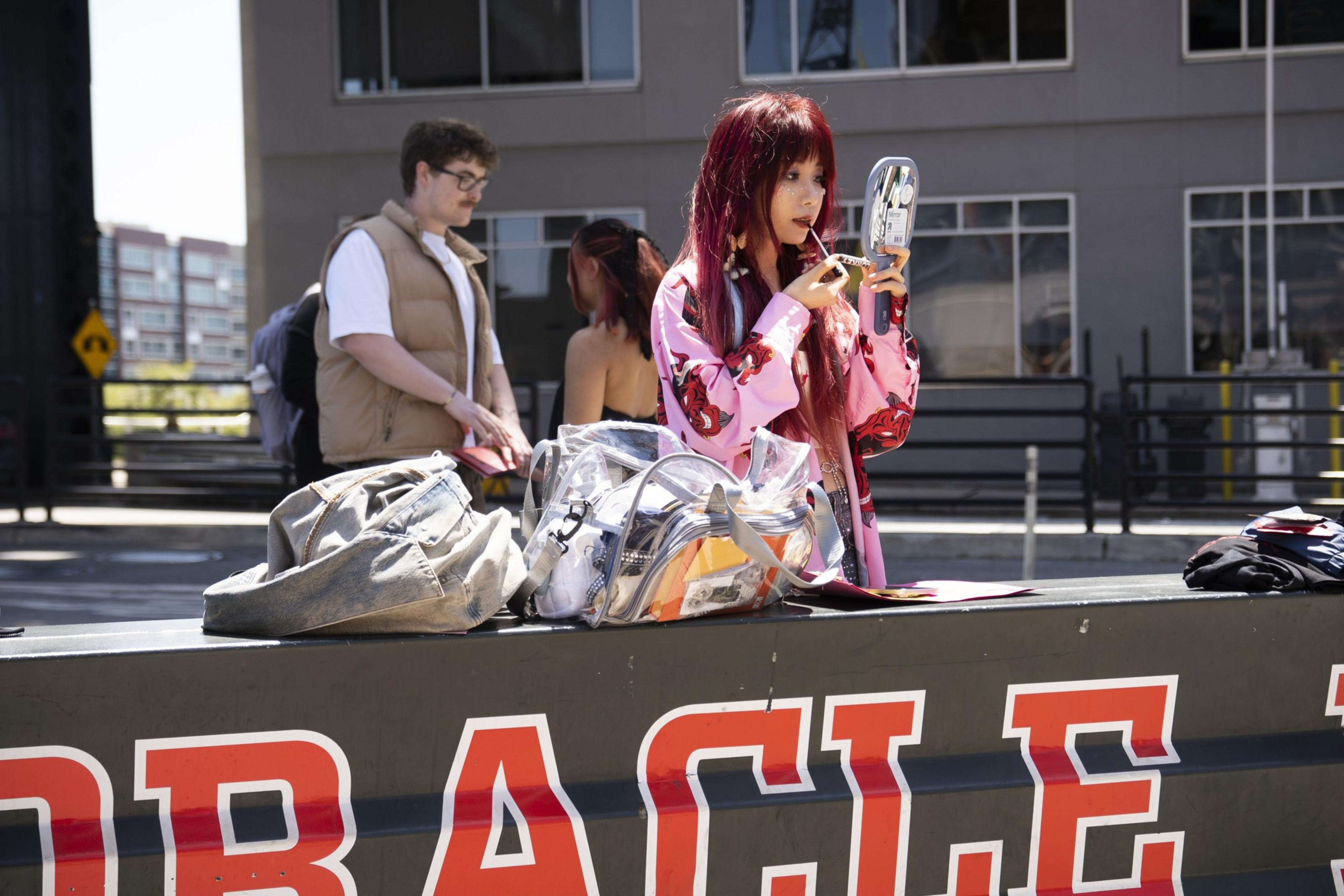 A person with long red hair in a pink outfit applies lipstick using a hand mirror. Nearby, a man stands, and bags are on a bench labeled &quot;ORACLE.&quot;