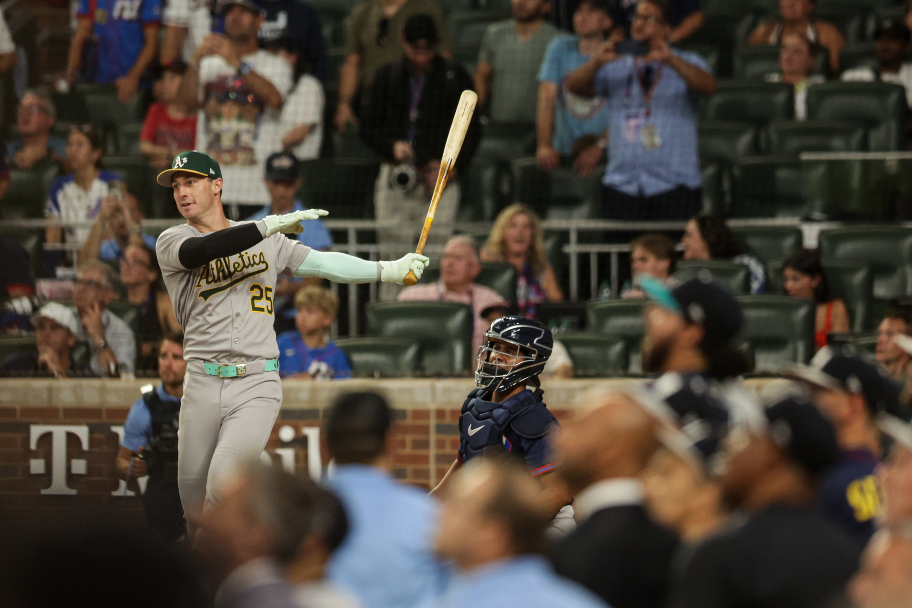 A baseball player in a gray uniform swings a bat, with a catcher behind him watching intently. Spectators fill the stadium seats in the background.