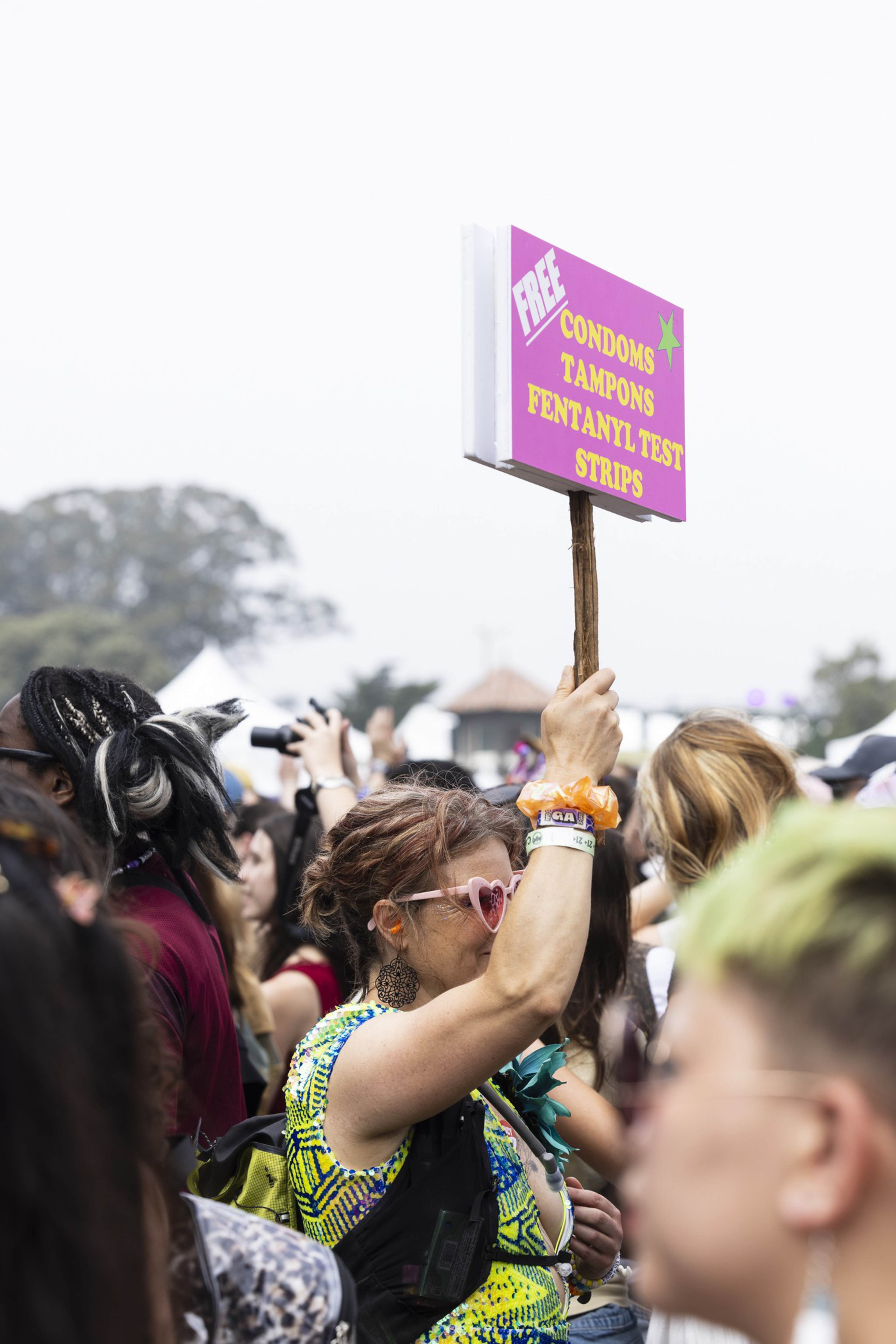 A person wearing heart-shaped sunglasses holds a sign reading "FREE CONDOMS TAMPONS FENTANYL TEST STRIPS" above a crowd at an outdoor event.