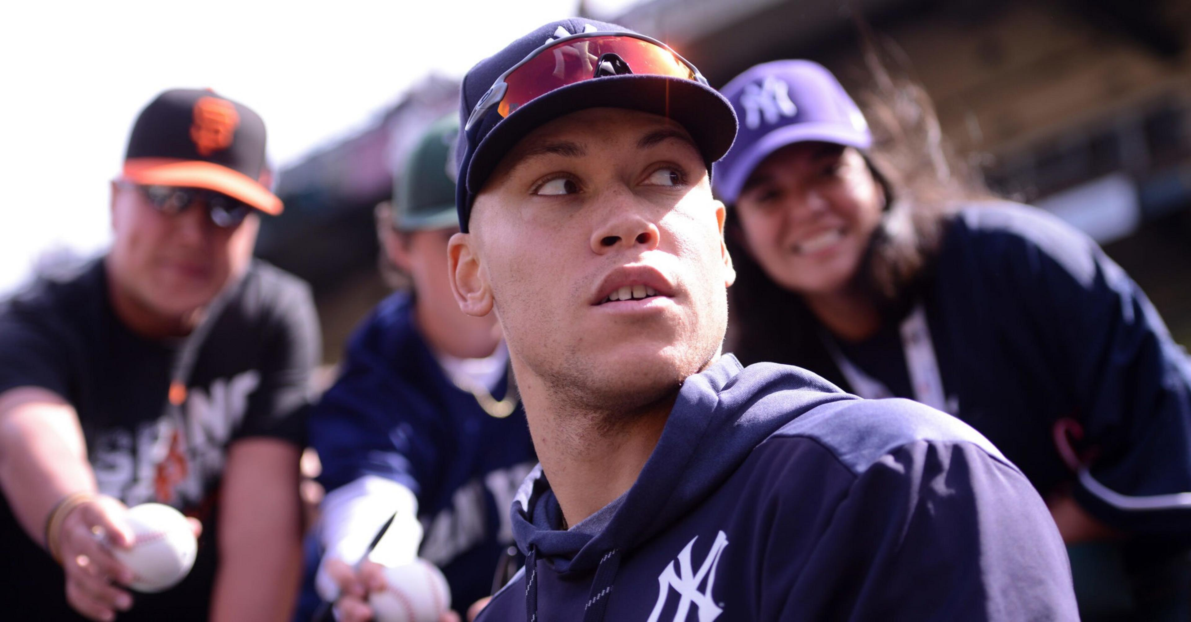 A baseball player in a New York Yankees cap and jacket looks to the side while fans behind him hold baseballs and wear team apparel.