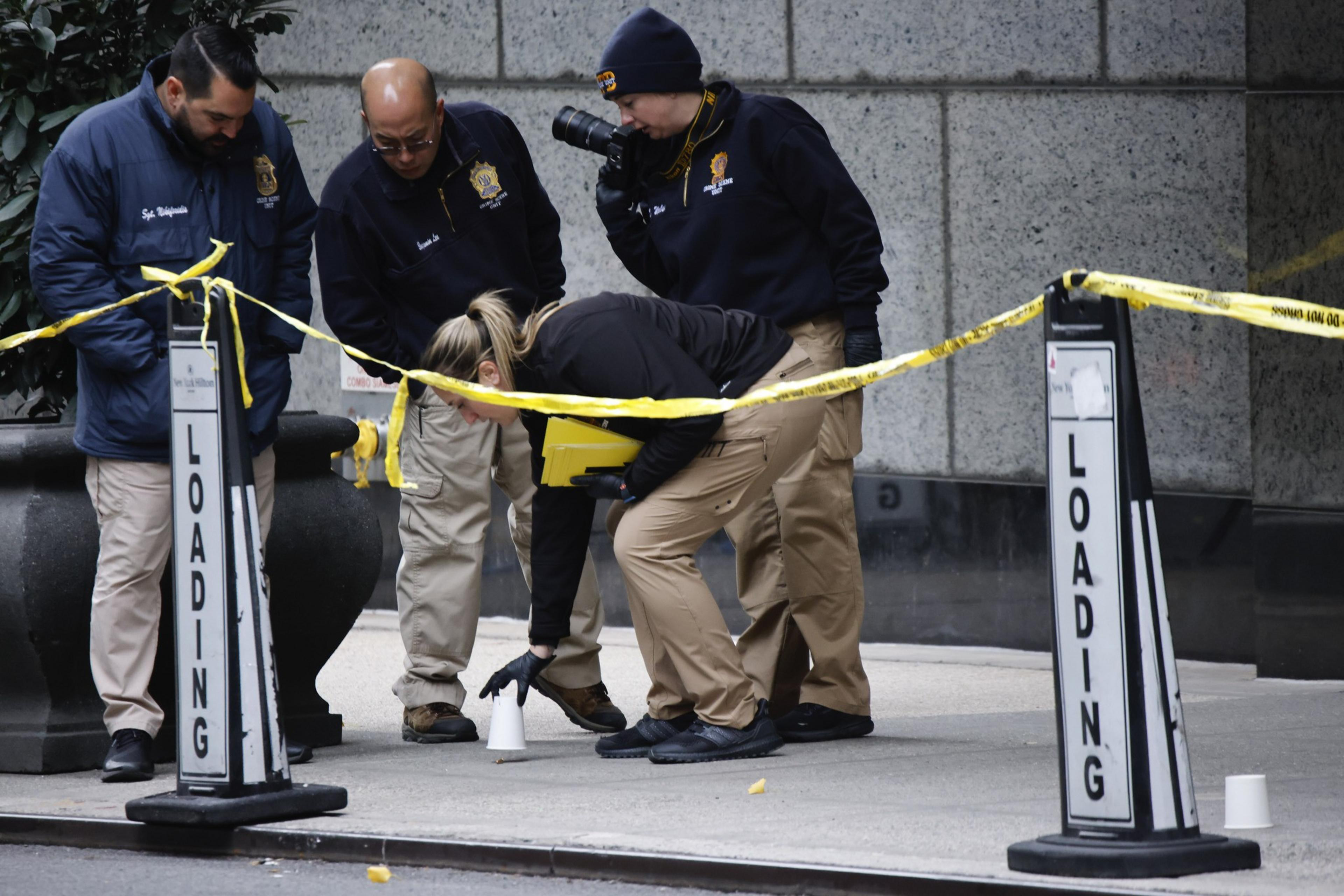 Four investigators examine a sidewalk, surrounded by yellow tape marked &quot;Loading.&quot; One crouches, placing a marker cone, while another takes photographs.
