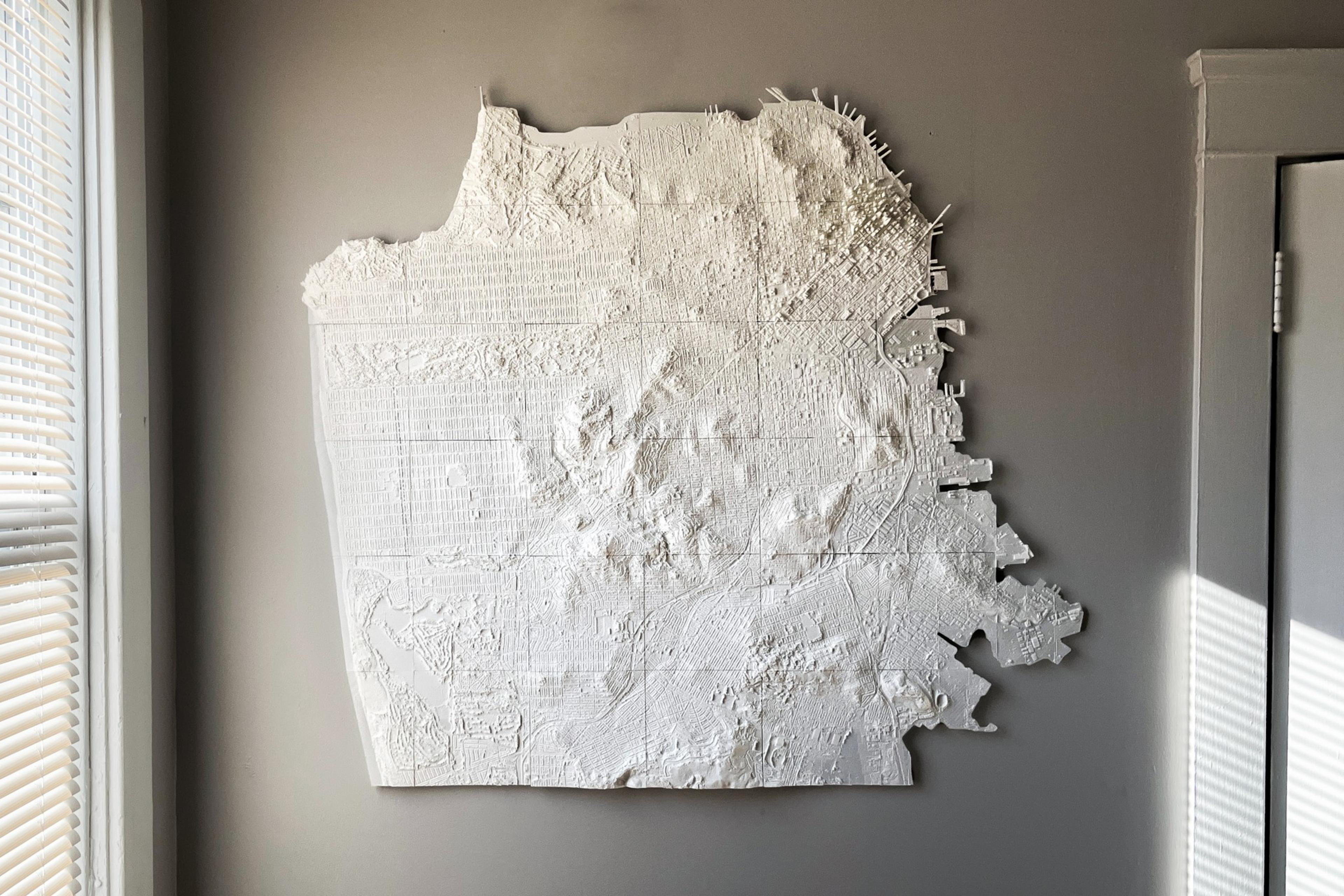 A textured white 3D relief map of a city with hills and streets hangs on a gray wall between a window with blinds and a door frame.