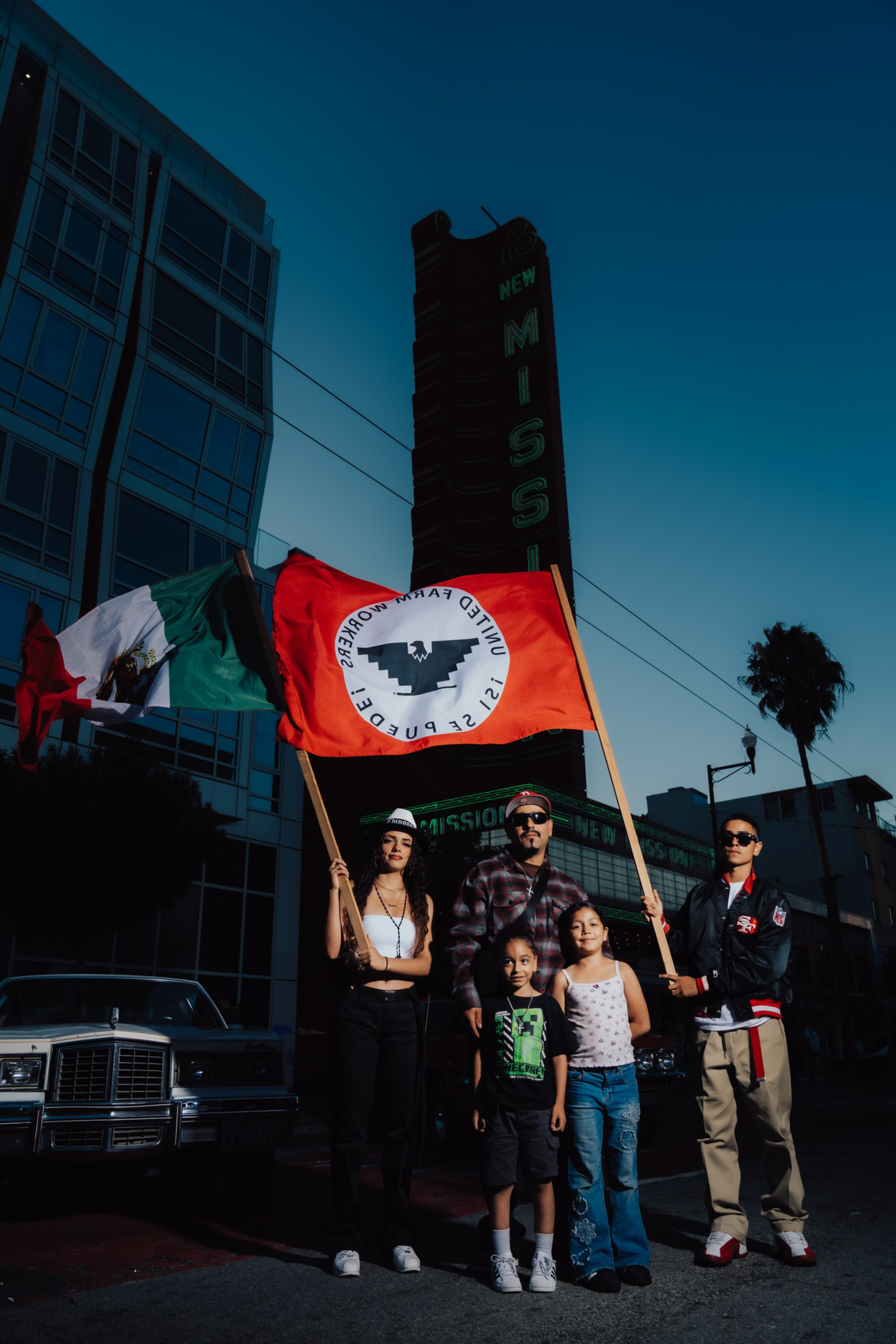 Five people stand on a street at dusk, holding a Mexican flag and a red flag with a black eagle and text, in front of a tall “New Mission” sign.