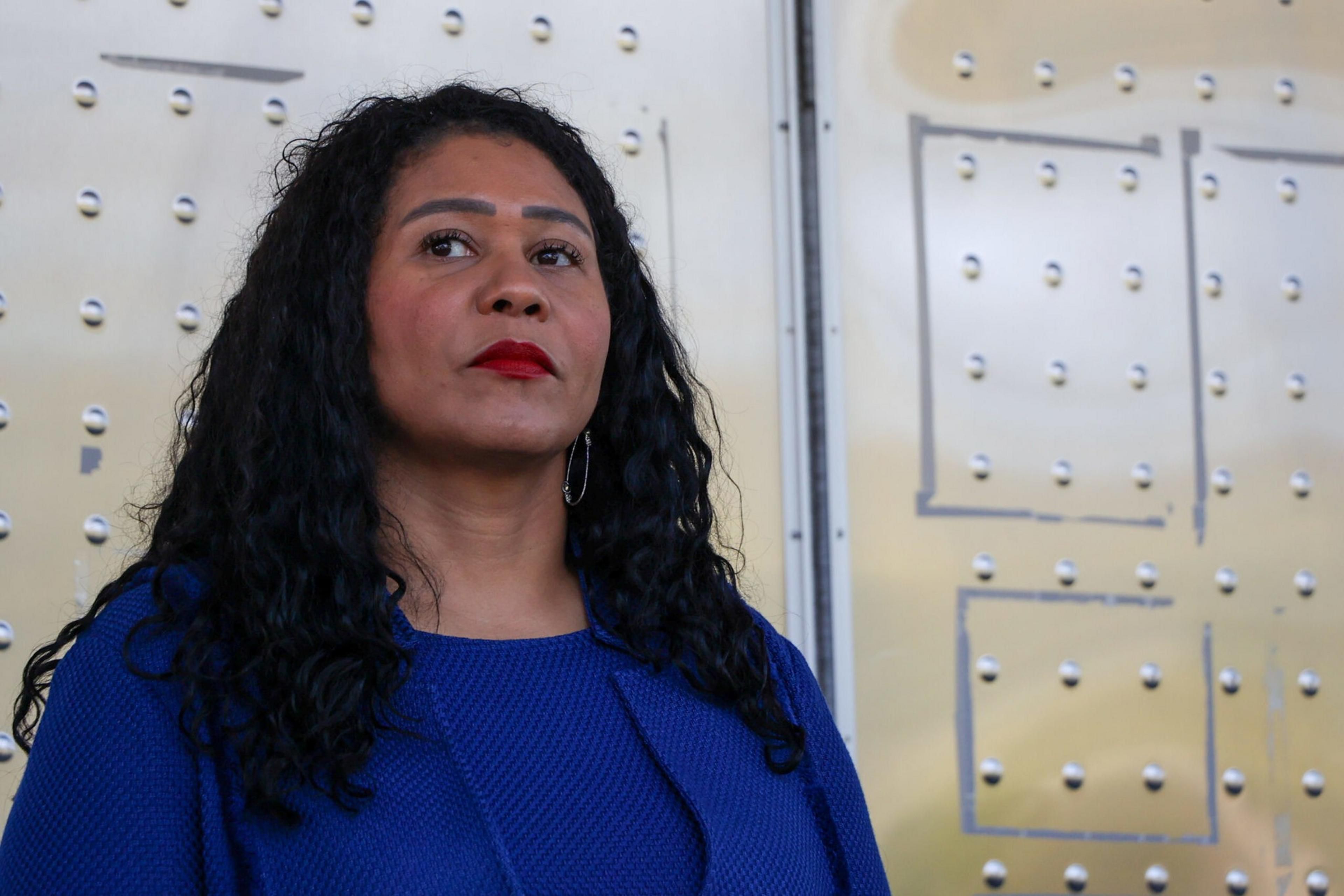 A woman with long curly black hair and red lipstick stands against a light-colored metal wall with rivets and square markings.