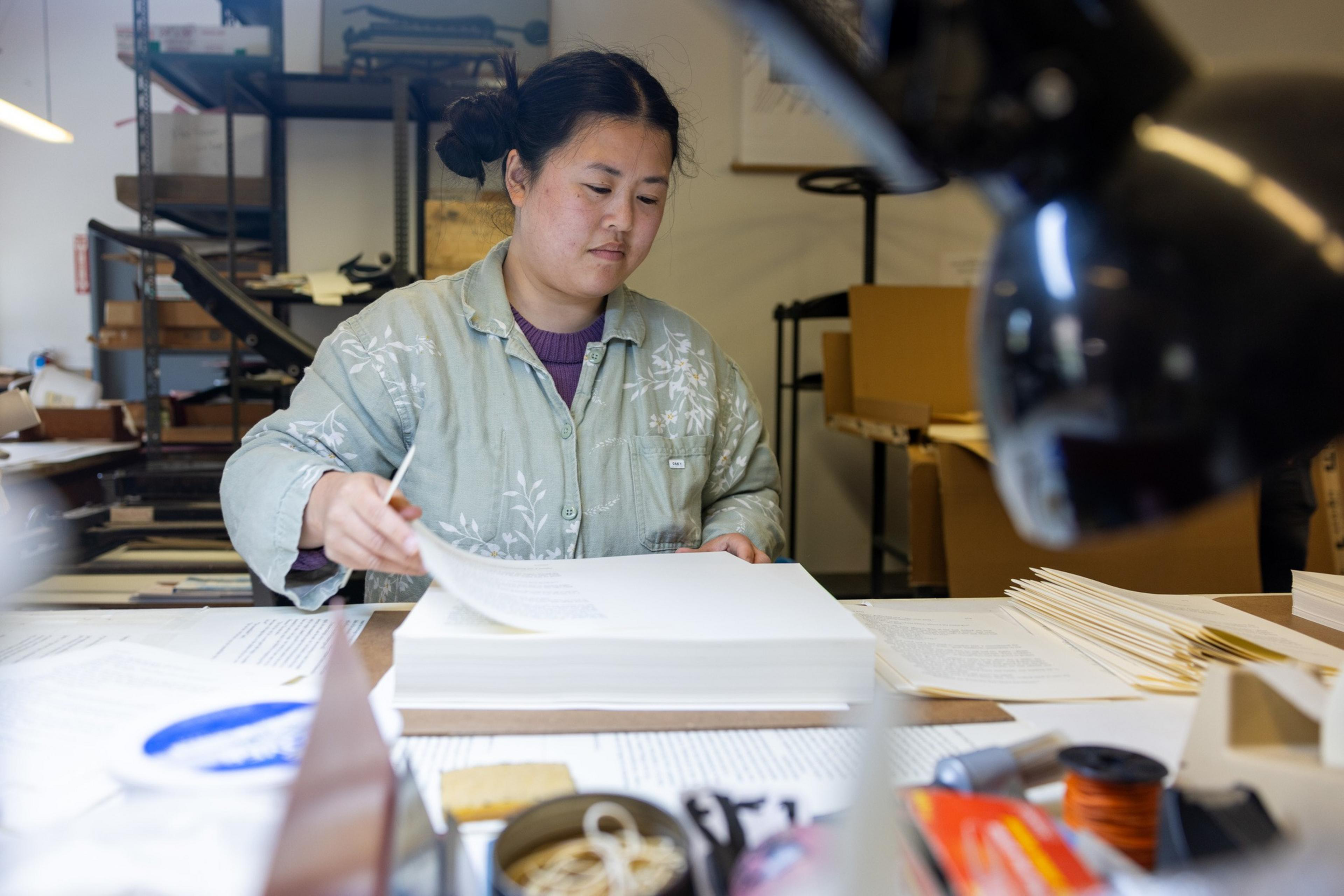 A woman in a workshop is inspecting a sheet of paper among stacks of documents on a desk cluttered with various items.
