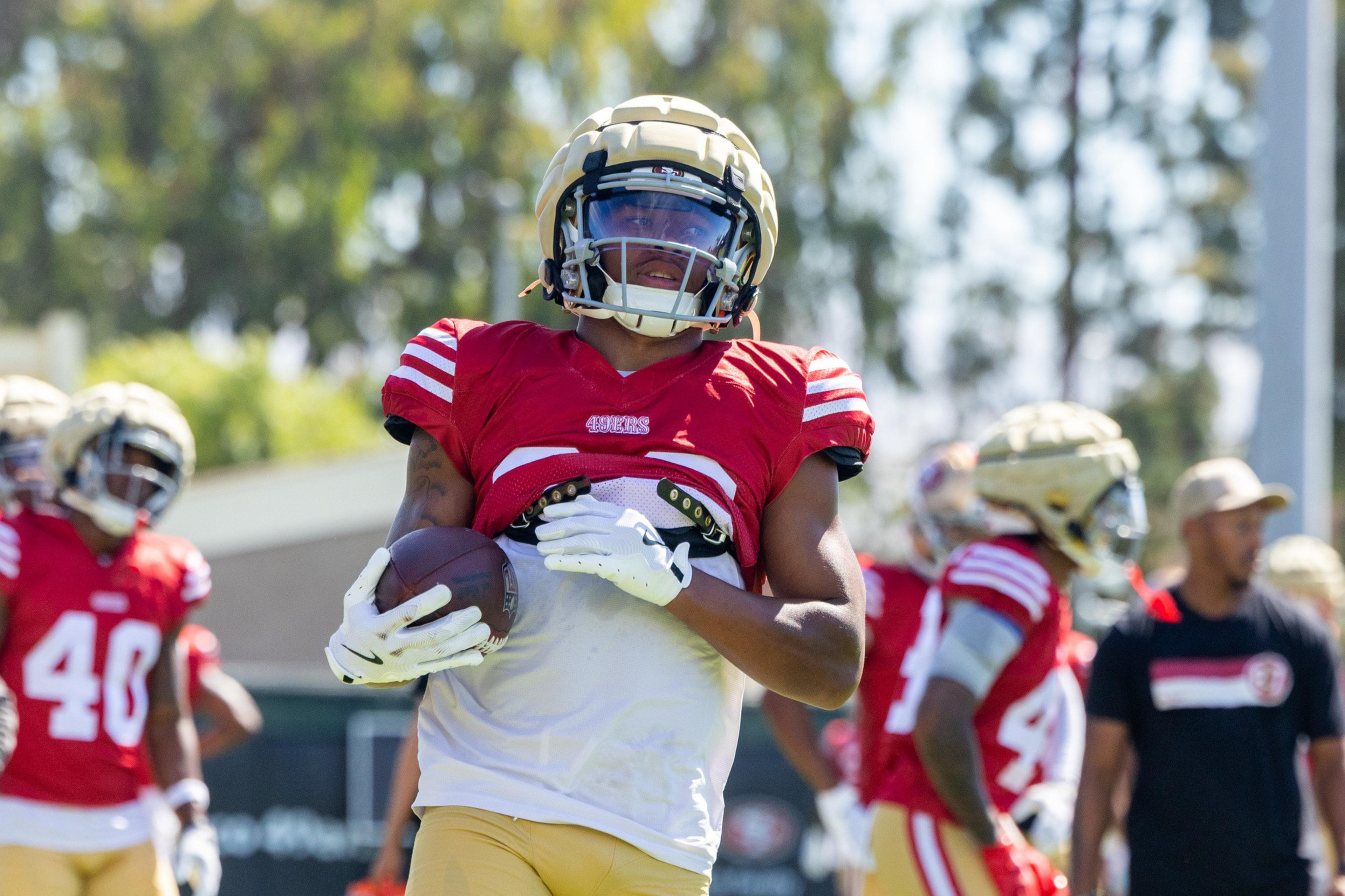 A football player in a red jersey and gold helmet clutches a football, with teammates and a coach in similar gear visible in the background.