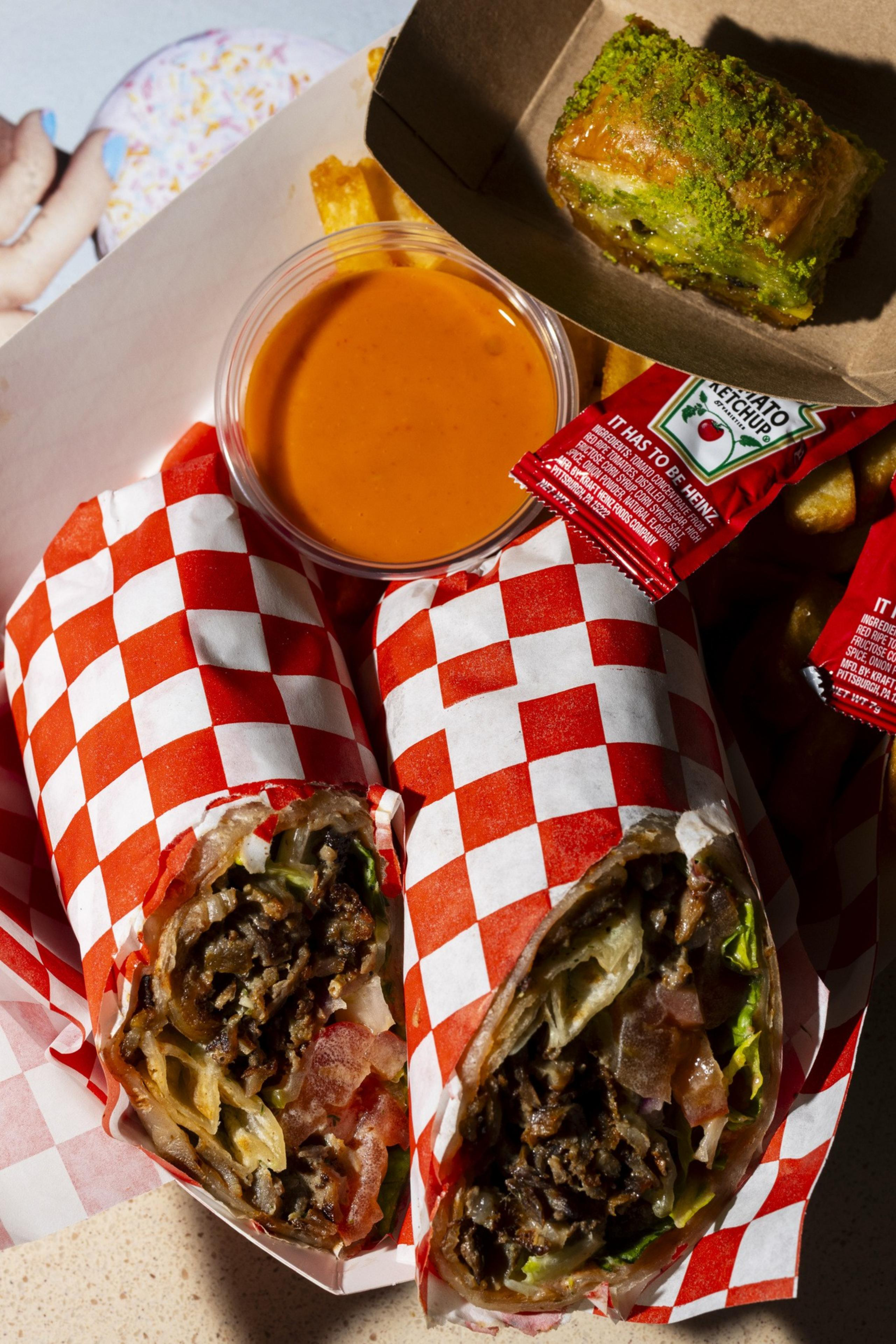 Two grilled wraps with meat and veggies are in red-checkered paper, accompanied by fries, ketchup packets, a container of orange sauce, and a pistachio dessert.