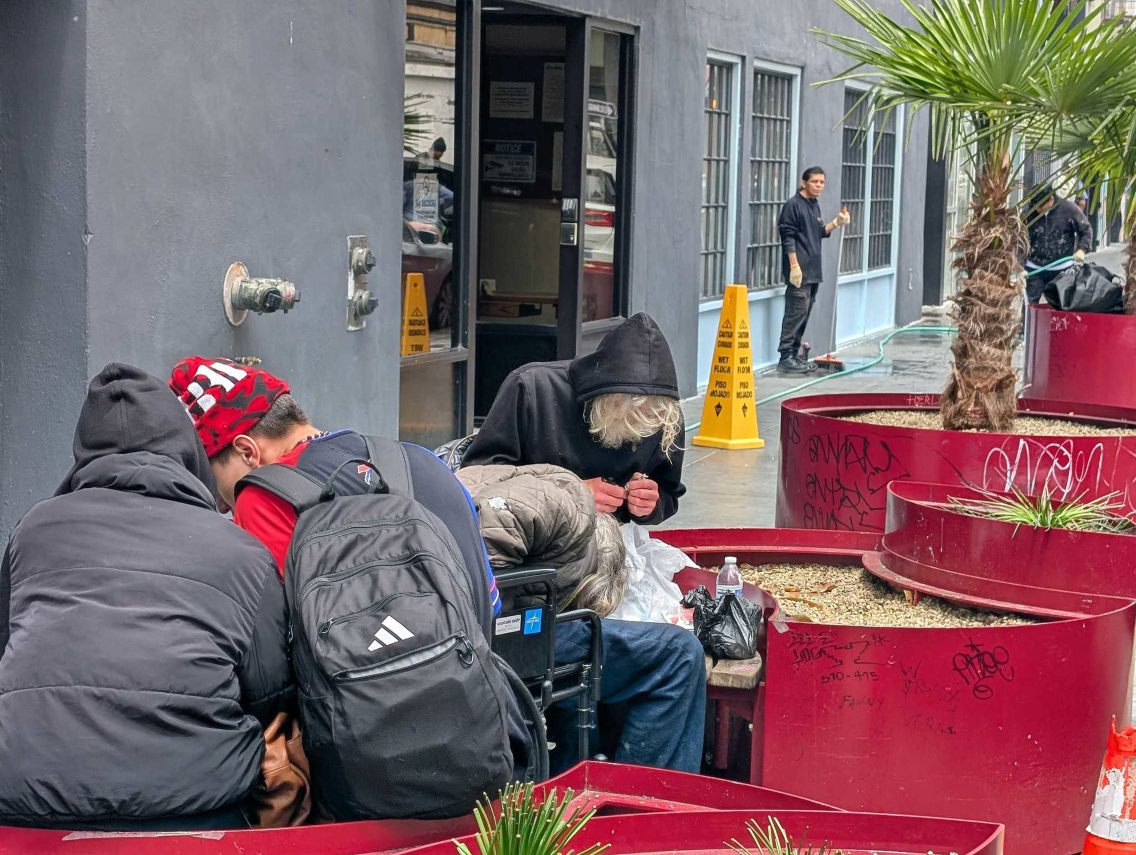 Three people in hoodies sit closely near red planters on a city sidewalk, with caution signs visible nearby. One person stands further away by a building entrance.