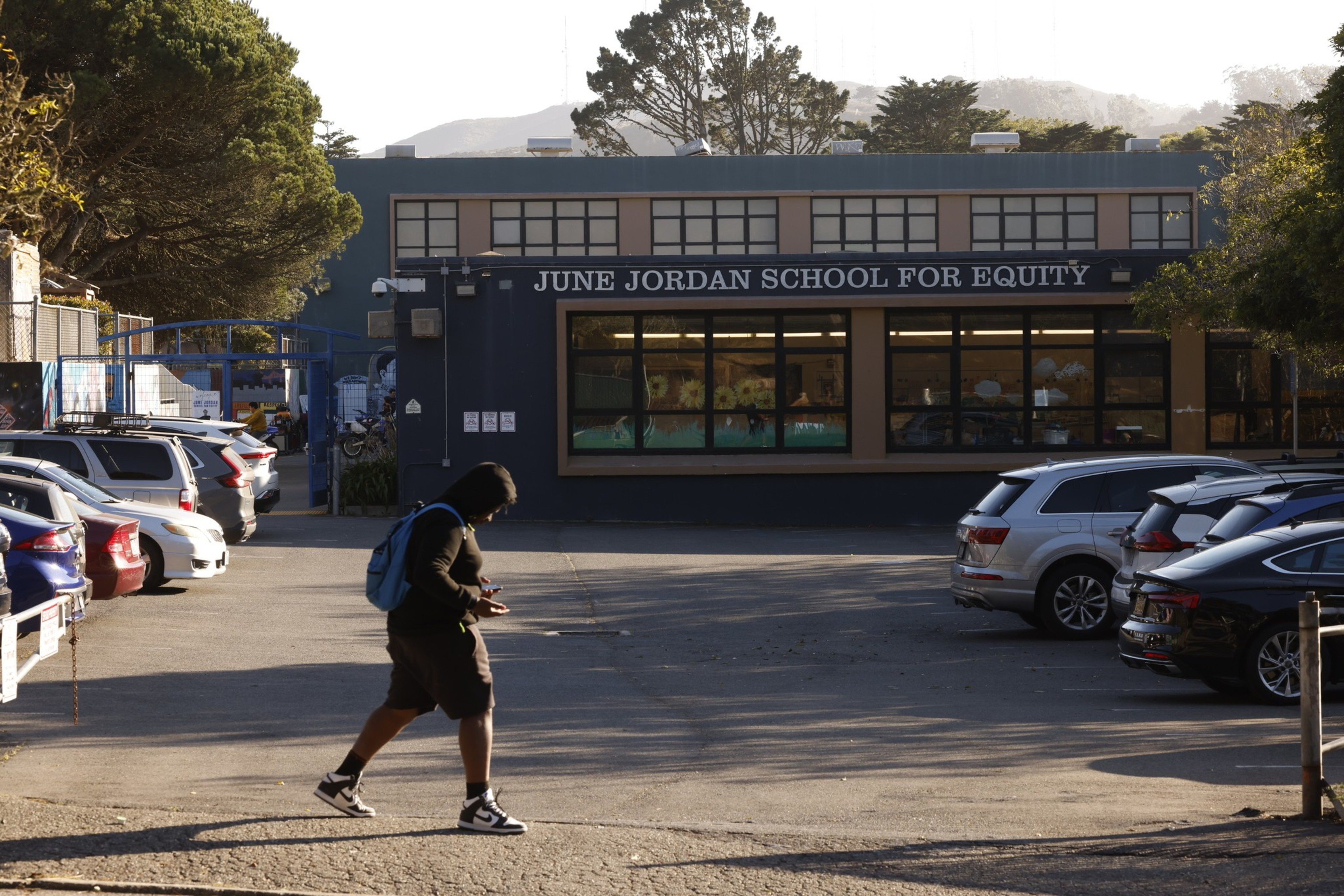 A person wearing a hoodie and backpack is looking at their phone while walking across a parking lot. The background shows a building labeled "June Jordan School for Equity."