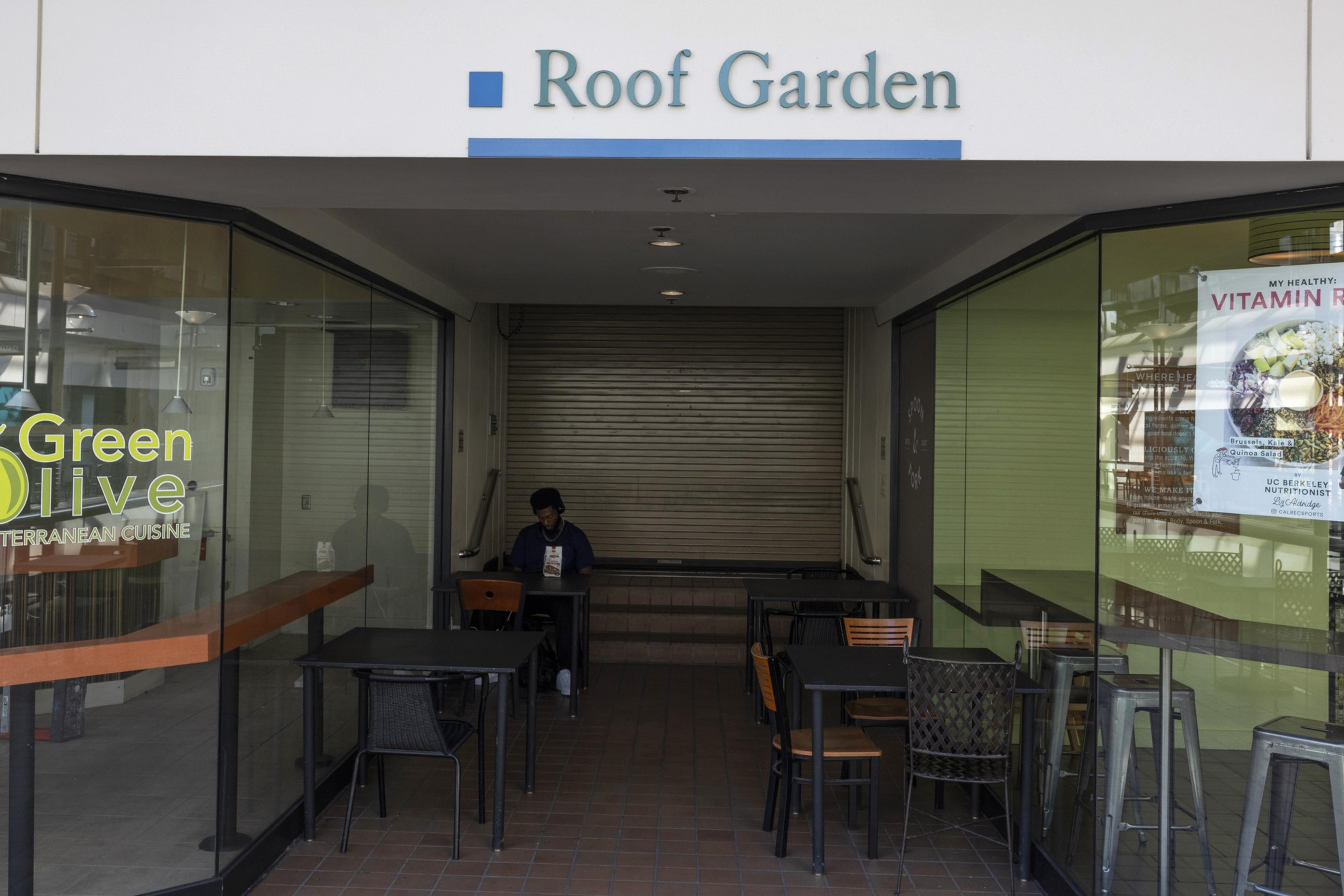 A small seating area with tables and chairs sits under a sign reading "Roof Garden," with glass walls and one person seated near a closed shutter.