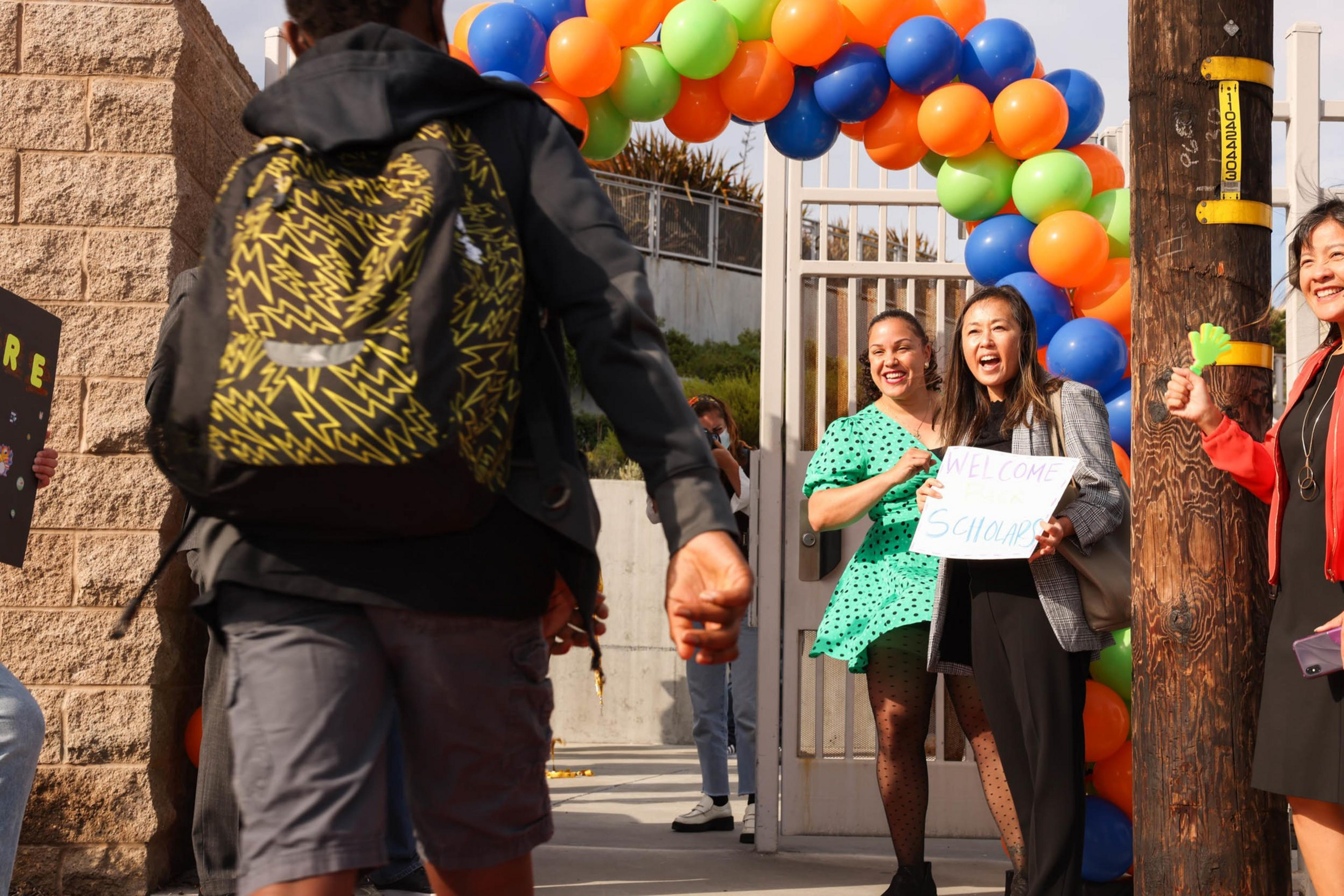 A student with a yellow and black backpack walks toward an entrance decorated with colorful balloons, greeted by smiling people holding welcome signs.
