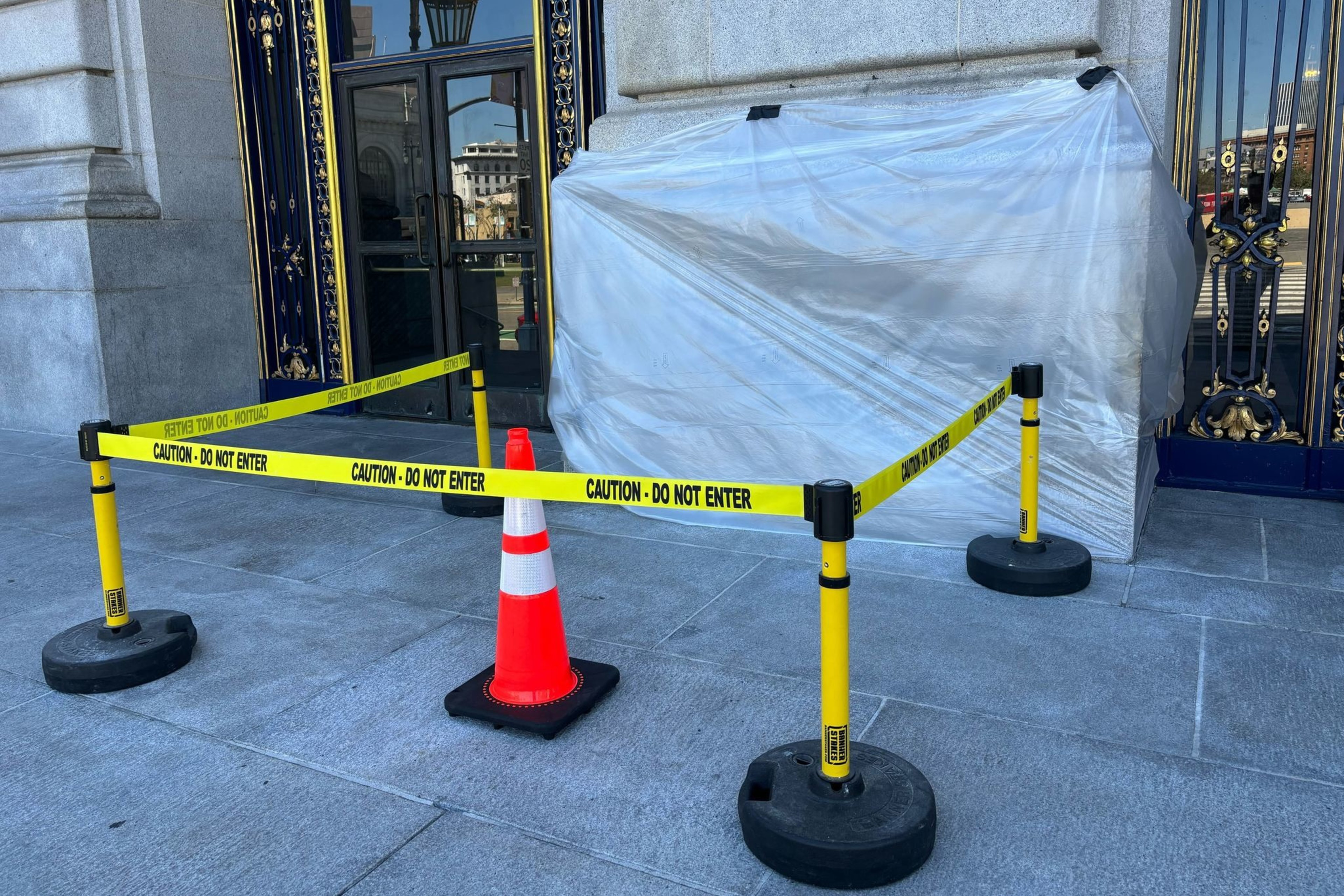 A doorway is blocked by "Caution - Do Not Enter" tape and a traffic cone. A large object covered in plastic is situated next to the door.